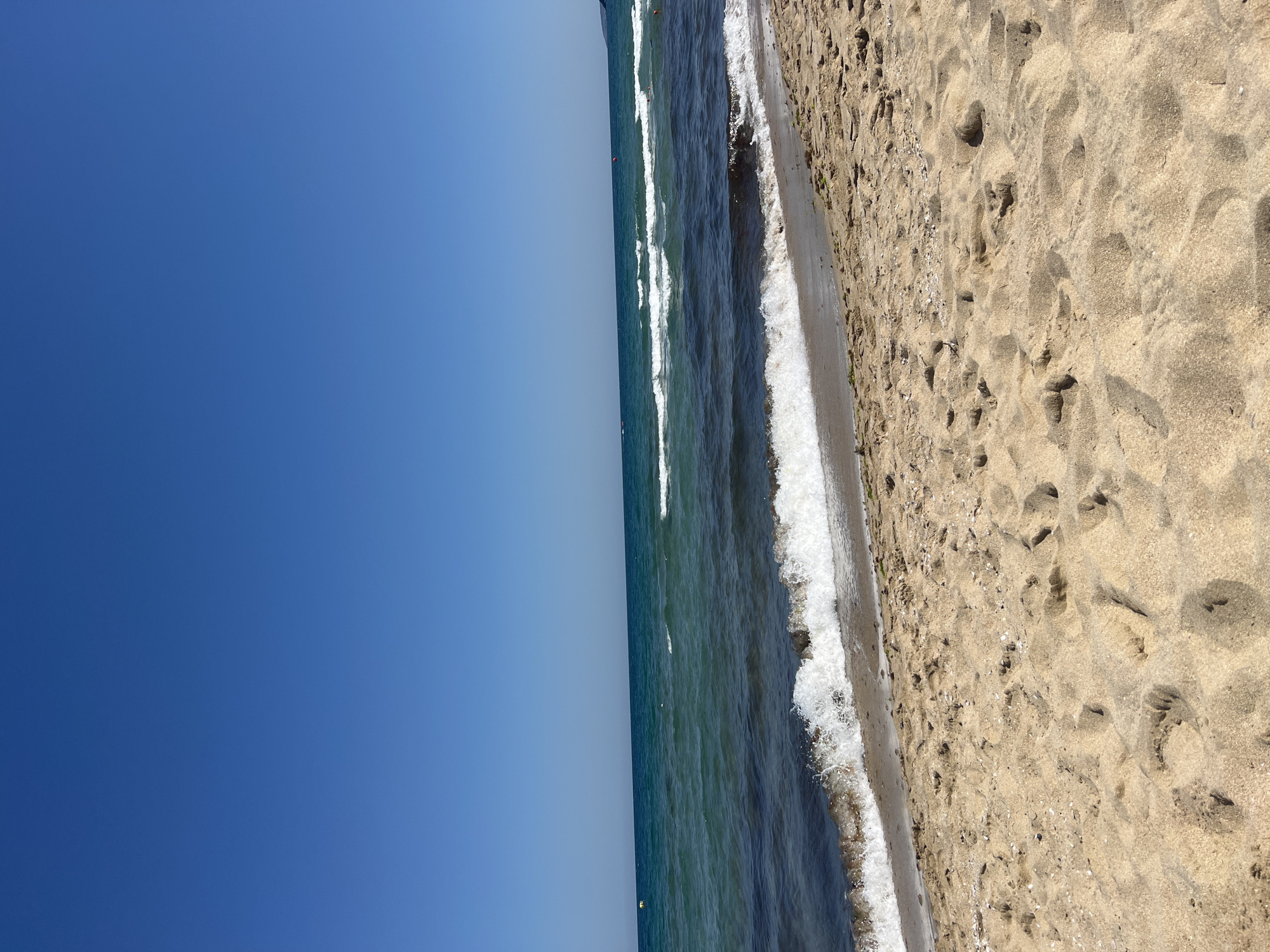 The image depicts a sandy beach with a clear blue sky overhead. The sea is a deep blue-green, with gentle waves breaking on the shore. The sand is light brown and appears to have some small shells and seaweed scattered on it. There are no people visible in the image. The scene is calm and tranquil. The location is Sozopol, Bulgaria.
