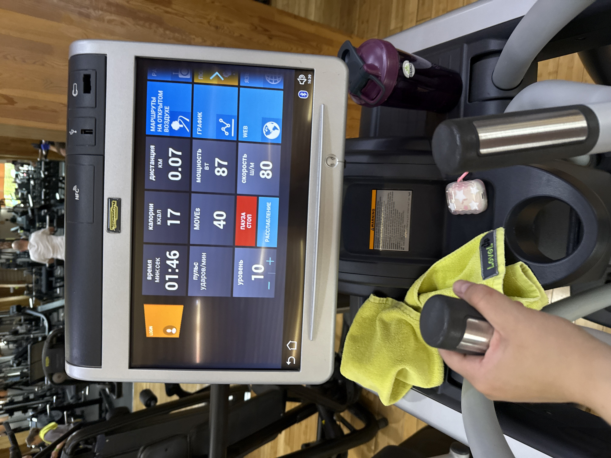 A close-up shot captures the console of a Technogym treadmill in a gym. The console screen displays workout statistics such as time (01:46), calories burned (17 kcal), distance (0.07 km), and speed (80 m/s). The screen also shows options in Russian, including "ROUTES IN THE OPEN AIR," "GRAPHIC," and "WEB." To the right of the console is a purple water bottle. A yellow "Level Up" towel drapes over the treadmill handle, which is being gripped by a person's hand. The surrounding gym has other exercise equipment visible in the background.
