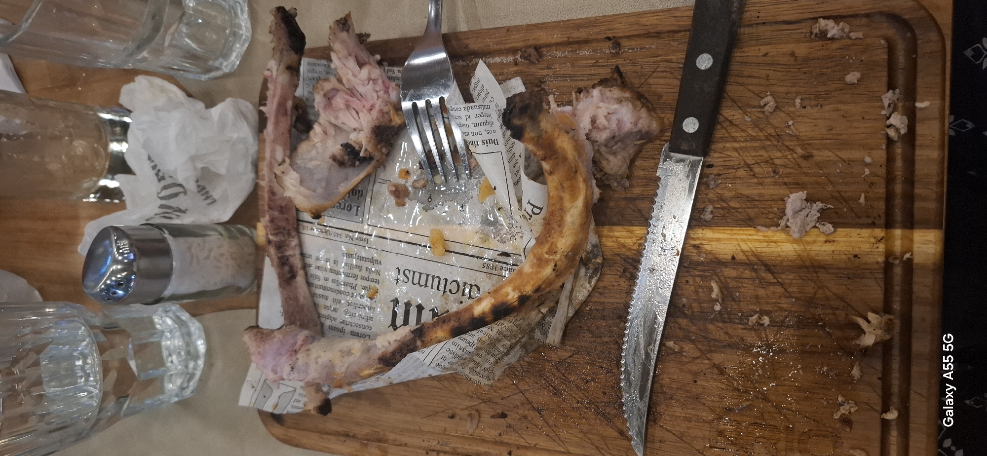 The image shows the remains of a meal on a wooden cutting board. There are several ribs with some meat still attached, a fork, a knife with a serrated blade, a salt shaker, and crumpled paper napkin. The ribs appear to be pork ribs. The cutting board is worn and has some juice stains. The background shows glasses and part of a table. The scene suggests that a meal has recently been finished. There is no specific information that can determine the location in the city of Yambol, Bulgaria.
