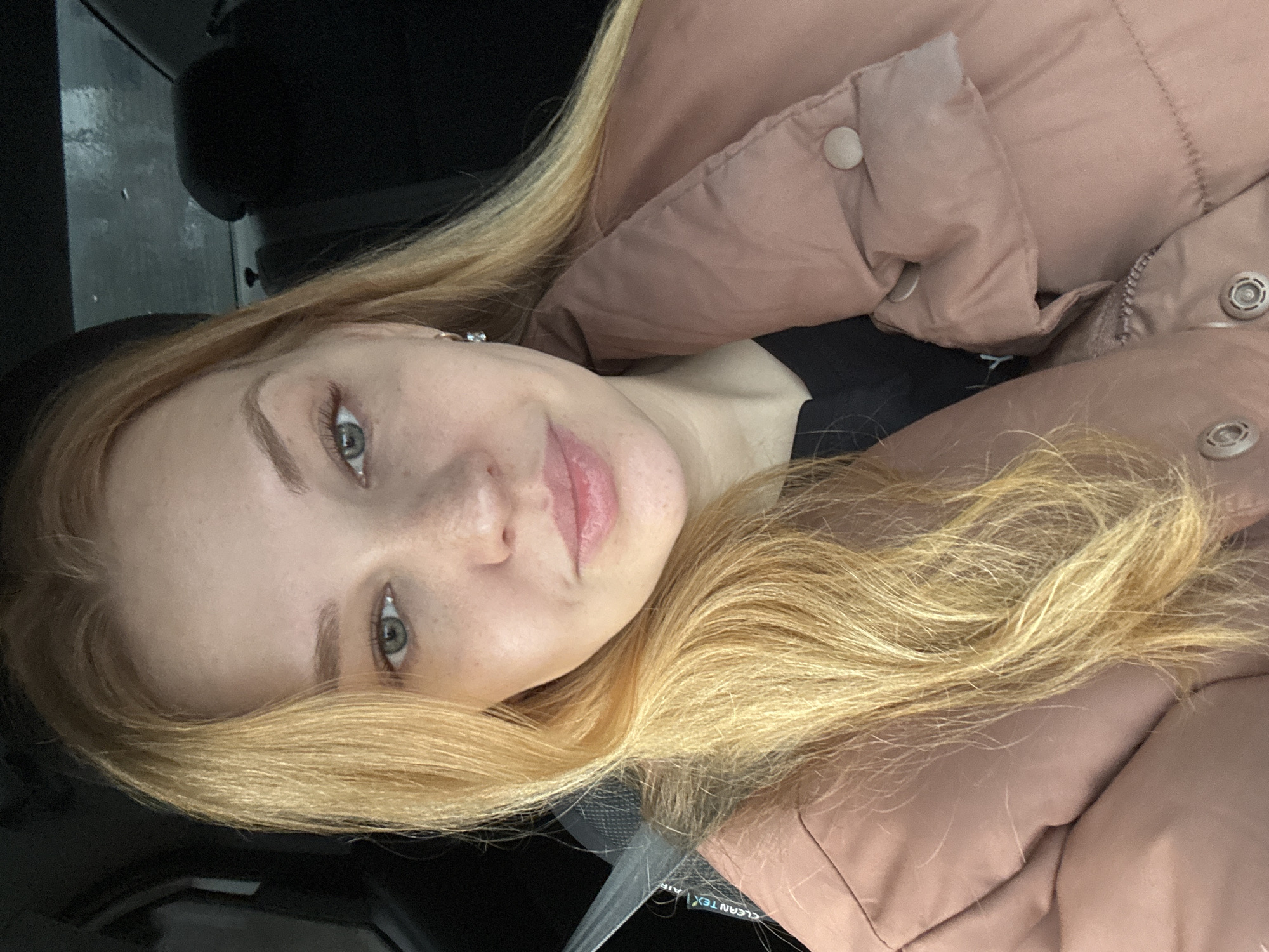 A young woman with light complexion and freckles is shown from a close-up, angled perspective. Her blond hair is long and wavy. She has grey eyes and light pink lips. She is wearing a black shirt and a pinkish-brown puffy jacket. A seatbelt is visible, indicating she is possibly in a car. The background is dark, suggesting a car interior. The location is not discernible from the image and cannot be confirmed to be Rusanovo, Russia.