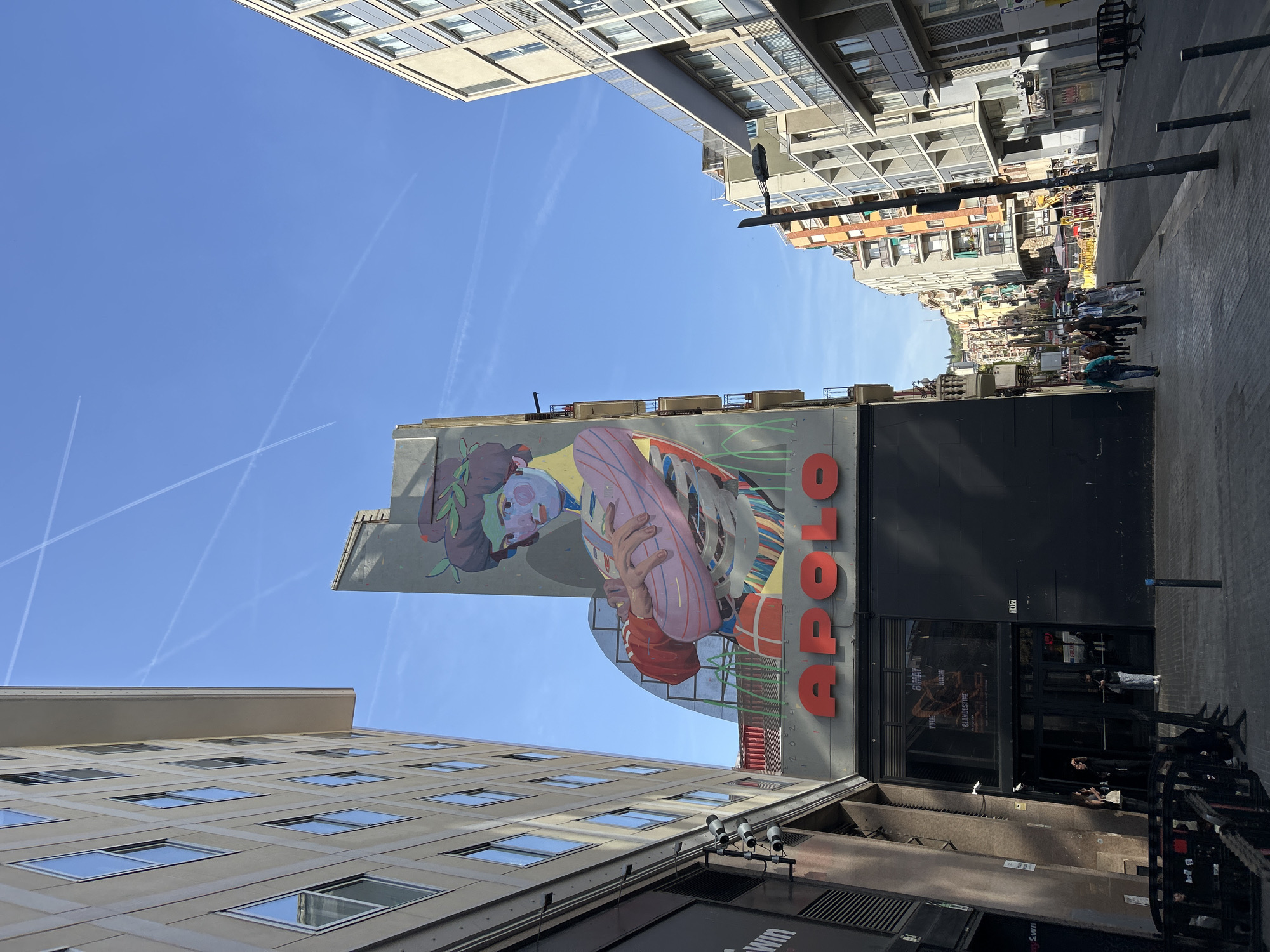 The image captures a vibrant urban scene, viewed from a low angle looking upwards, showcasing a narrow street lined with buildings under a clear blue sky. On the right, a large, colorful mural adorns the side of a building, depicting an abstract figure with a green, leafy headpiece holding a pink, striped object. Below the mural, a prominent red sign reads "APOLO". Further down, a dark storefront is visible, reflecting the surroundings and containing faint, unreadable text inside.
On the left, a modern building with a grid of square windows rises towards the sky. On the opposite side, a mix of buildings with balconies and varied facades extends into the distance. Several pedestrians are visible walking along the sidewalk in the background, suggesting a lively street. The sky is bright blue, crisscrossed by multiple white airplane contrails, indicating clear and sunny weather. The time of day appears to be mid-day due to the bright, direct sunlight. The visible "APOLO" sign is a strong location cue for Barcelona, Spain, specifically referencing the famous Sala Apolo music venue. The scene depicts everyday urban activity, with people going about their day in a bustling city environment.