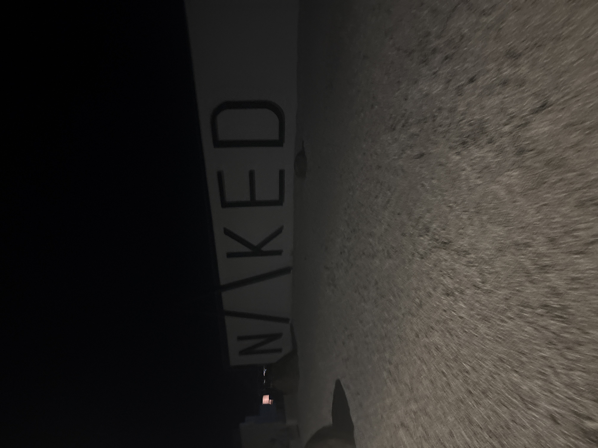 The image depicts a nighttime scene outdoors in Burgas, Bulgaria. A large, dark sign with the word "NAKED" in white capital letters is mounted on a light-colored wall. The letters are stylized, with the "N" followed by a slanting line, then "KED." The ground in front of the wall appears to be a sandy or gravelly surface, and there is some indistinct debris or small mounds on it. To the left of the sign, there are some structures, possibly artificial and illuminated from within, as one appears to have a soft orange glow. The overall lighting is dim, indicating it is nighttime, with artificial lights providing illumination. The camera perspective is low, looking up towards the sign and wall. There are no people clearly visible in the image.