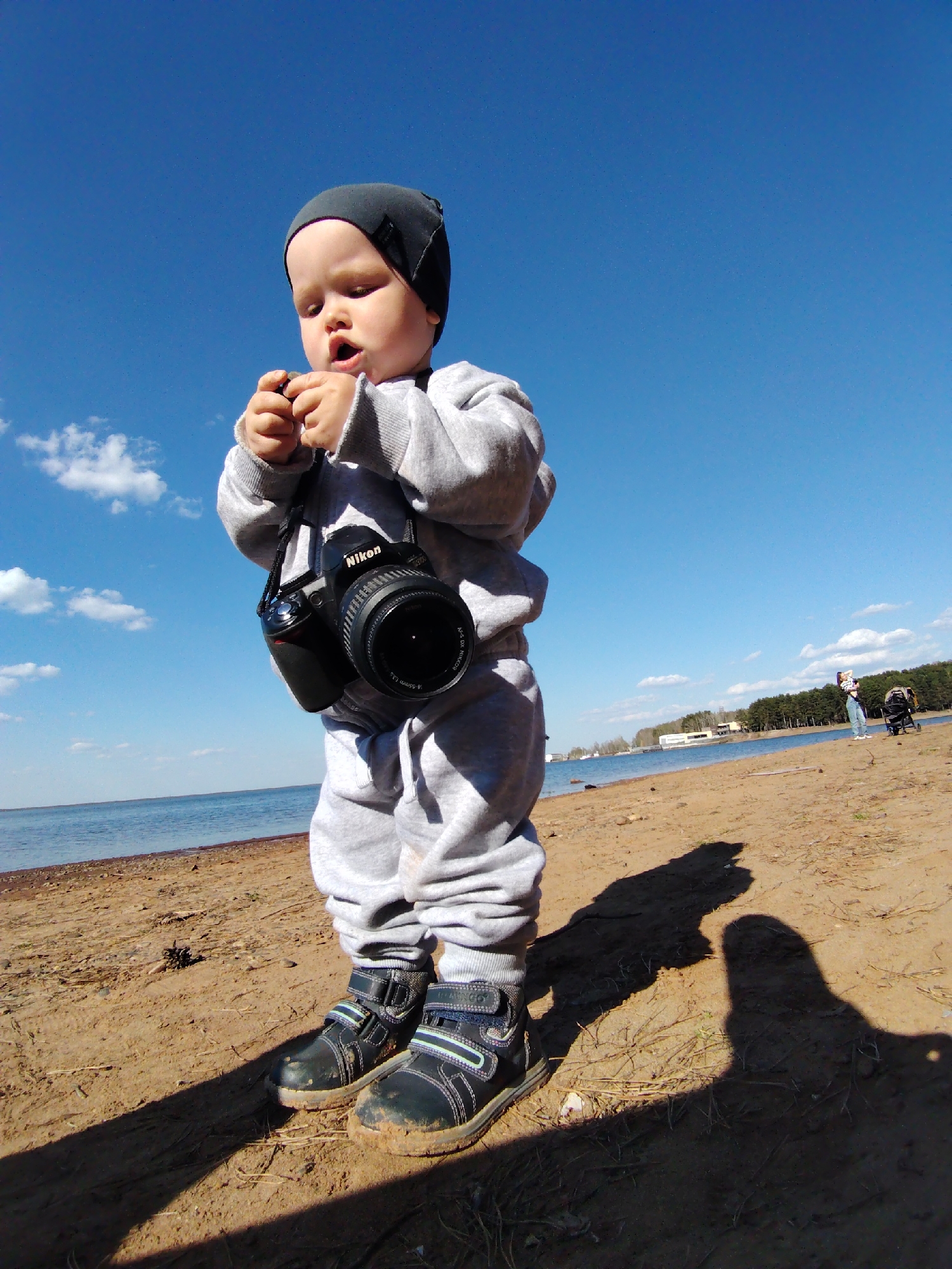 A toddler stands on a sandy beach wearing a gray tracksuit, black hat, and dark-colored boots that are covered in mud. A black Nikon camera hangs around their neck. The child is holding a small object in their hands and has an open-mouthed expression. The sky is clear blue with scattered clouds. In the background, there is a body of water and a distant shoreline with trees and buildings. Further in the background, two individuals and a stroller are seen. The location is likely the beach area in Chaikovsky, Russia, due to the environment and location of the town on the Kama River. A shadow of an adult figure is visible on the right side of the frame, suggesting the picture was taken by them.