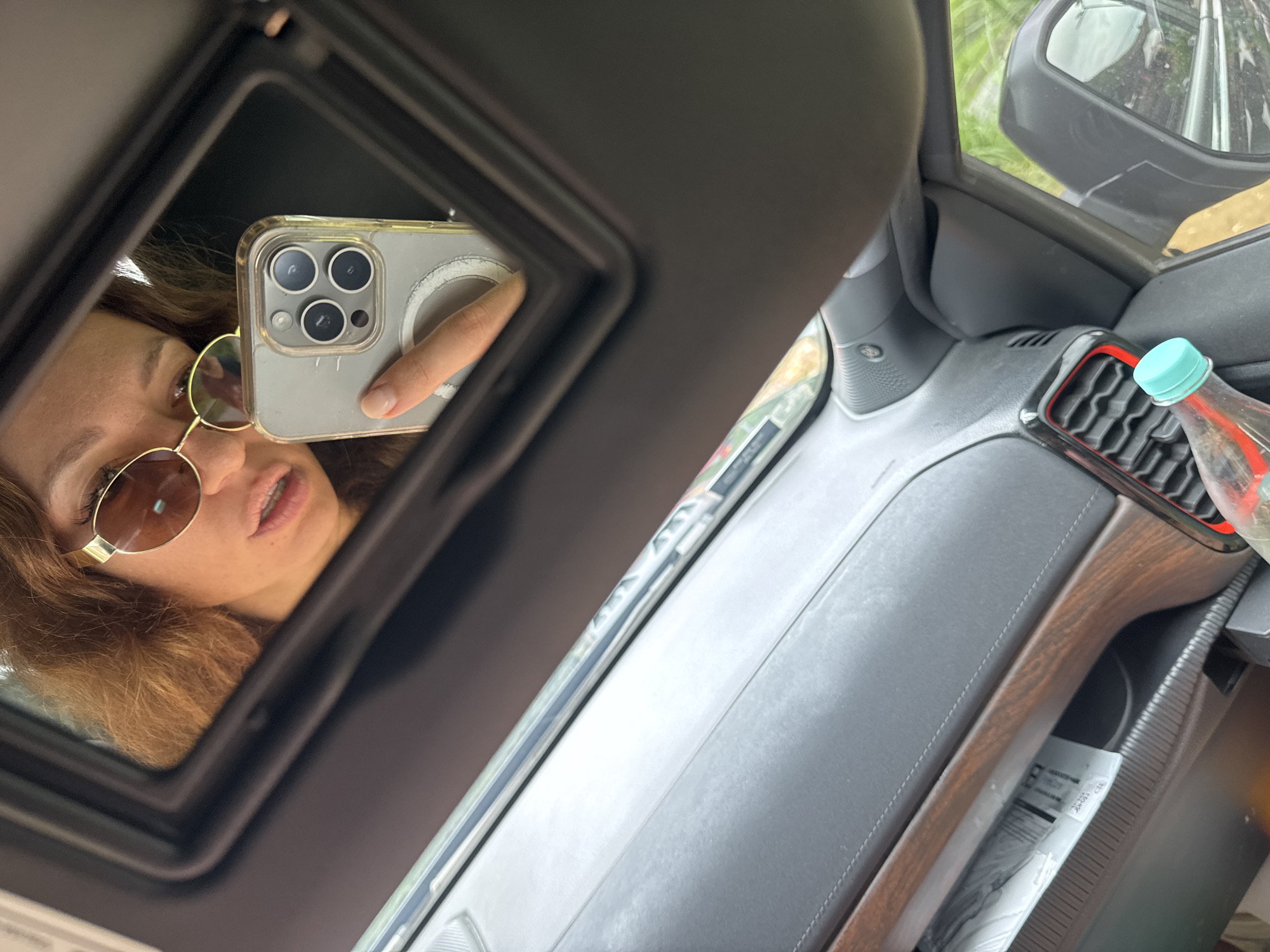 The scene shows the interior of a vehicle, likely a car, with a woman taking a selfie in the vanity mirror of the sun visor. The woman has curly hair and is wearing sunglasses. She is holding a smartphone with three camera lenses and a clear case. The car's dashboard is visible, with a water bottle in a holder near the air vents. Part of the car's side mirror is also visible. The location of Yên Minh Commune, Vietnam cannot be determined from this image.