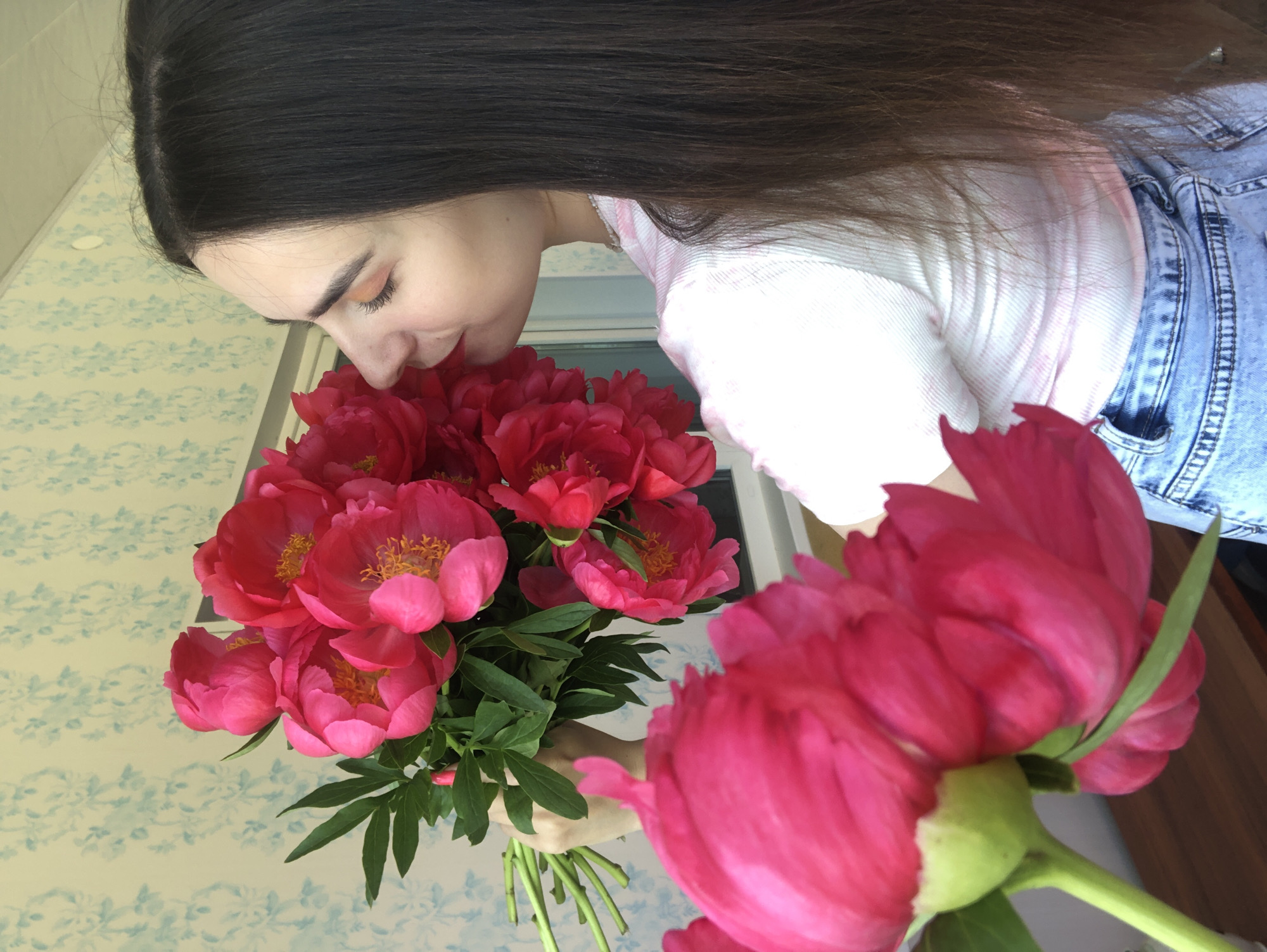 A young woman with long, dark hair is holding a bouquet of bright pink peonies and smelling them. She is wearing a pink and white striped t-shirt and blue denim shorts. The background is a wall with a light blue floral wallpaper. The image appears to be taken indoors, possibly in a home, as suggested by the wallpaper and wooden surface in the foreground. No specific location in Noviy, Russia is identifiable from the image.