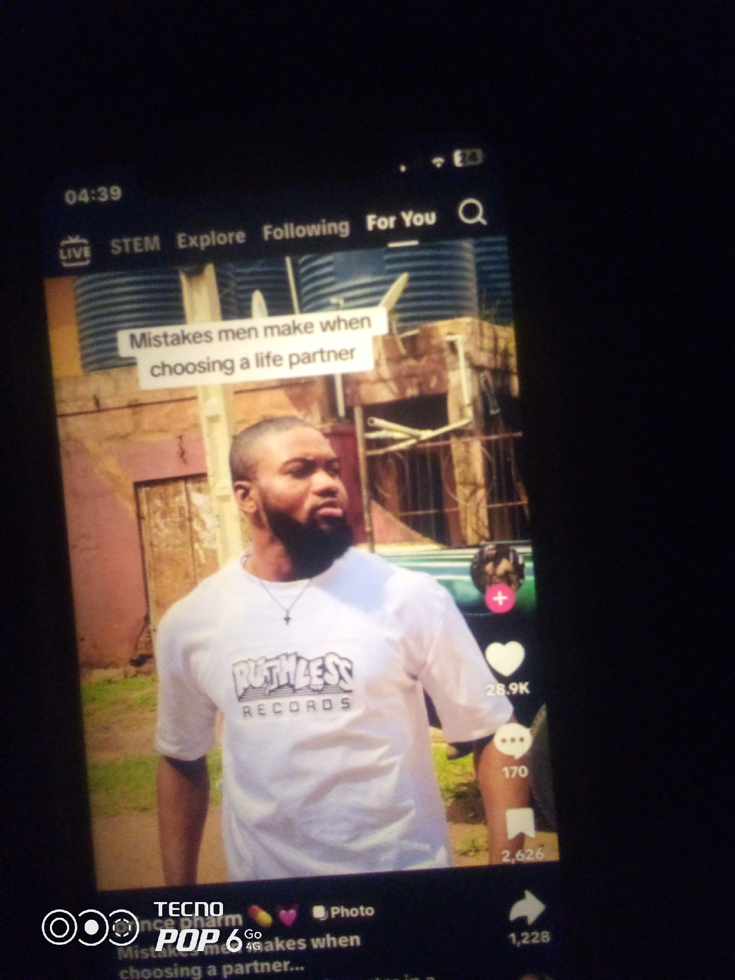 The image is a screenshot of a mobile phone displaying a social media video. The video features a man with a beard, wearing a white t-shirt with "RUTHLESS RECORDS" printed on it. He is looking to his right. Text overlays on the video read "Mistakes men make when choosing a life partner". The background appears to be an urban environment with buildings and what look like water tanks. The bottom of the screen shows icons and text indicating it's a TECNO POP 6 phone, with options like "Photo", a heart icon with "28.9K", a speech bubble icon with "170", a bookmark icon with "2,626", and a share icon with "1,228". The time "04:39" is visible in the top left corner, along with signal and battery indicators. The specific location in the city of Howayi, Nigeria cannot be determined from the image.