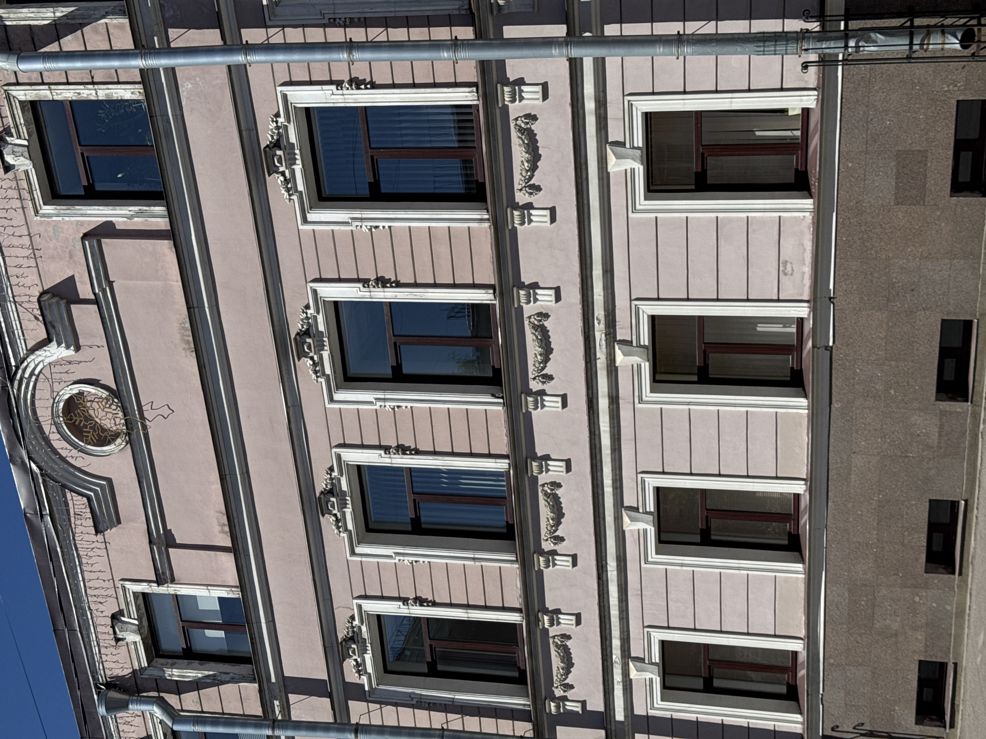The image displays the facade of a multi-story building located at Nevsky Prospect, 61, Saint Petersburg, Russia. The building features a pale pink or dusty rose main facade adorned with white architectural trim and dark brown window frames, illuminated by direct sunlight under a clear blue sky.
The uppermost visible story includes a central, recessed circular medallion containing an ornate bronze or gold-colored metal snowflake ornament. Below the roofline, white decorative molding is visible, with strands of festive-style string lights (resembling icicles) hanging from it. A single rectangular window with a dark brown frame and an elaborate white surround is visible on the far left of this level. A silver-colored downspout runs vertically along the far left edge of the building.
The two middle stories feature repeating sets of rectangular windows, each with dark brown frames and intricate white decorative surrounds, including pediments and pilasters. Between these windows, the pink facade is horizontally striated, and ornate white relief garlands and smaller rectangular pilaster-like elements are positioned.
The ground story is characterized by a dark brown, coarse-textured granite-like facade that extends up to the window sills. Above the granite, the pale pink facade continues with horizontally striated detailing. Rectangular windows with dark brown frames and simpler white pilaster-and-entablature surrounds are present. At street level, several small, recessed rectangular basement windows with dark frames are visible. A silver-colored downspout runs vertically along the far right side of the building, with its lower section encased within a protective black metal fence or railing. No people are visible in the scene.