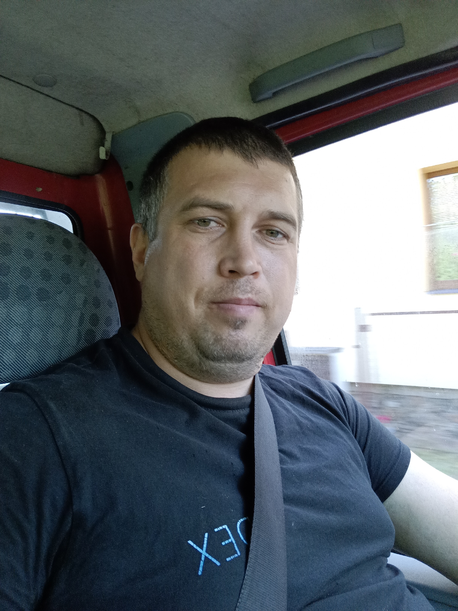 A man with short brown hair and a light beard is seated in a vehicle, looking directly at the camera. He is wearing a black t-shirt with a blue graphic on it and a seatbelt. The interior of the vehicle is visible, including a red panel on the left and a grey seat. Outside the vehicle, a white building with a window is visible. The image was taken in Dalovy, Czechia.