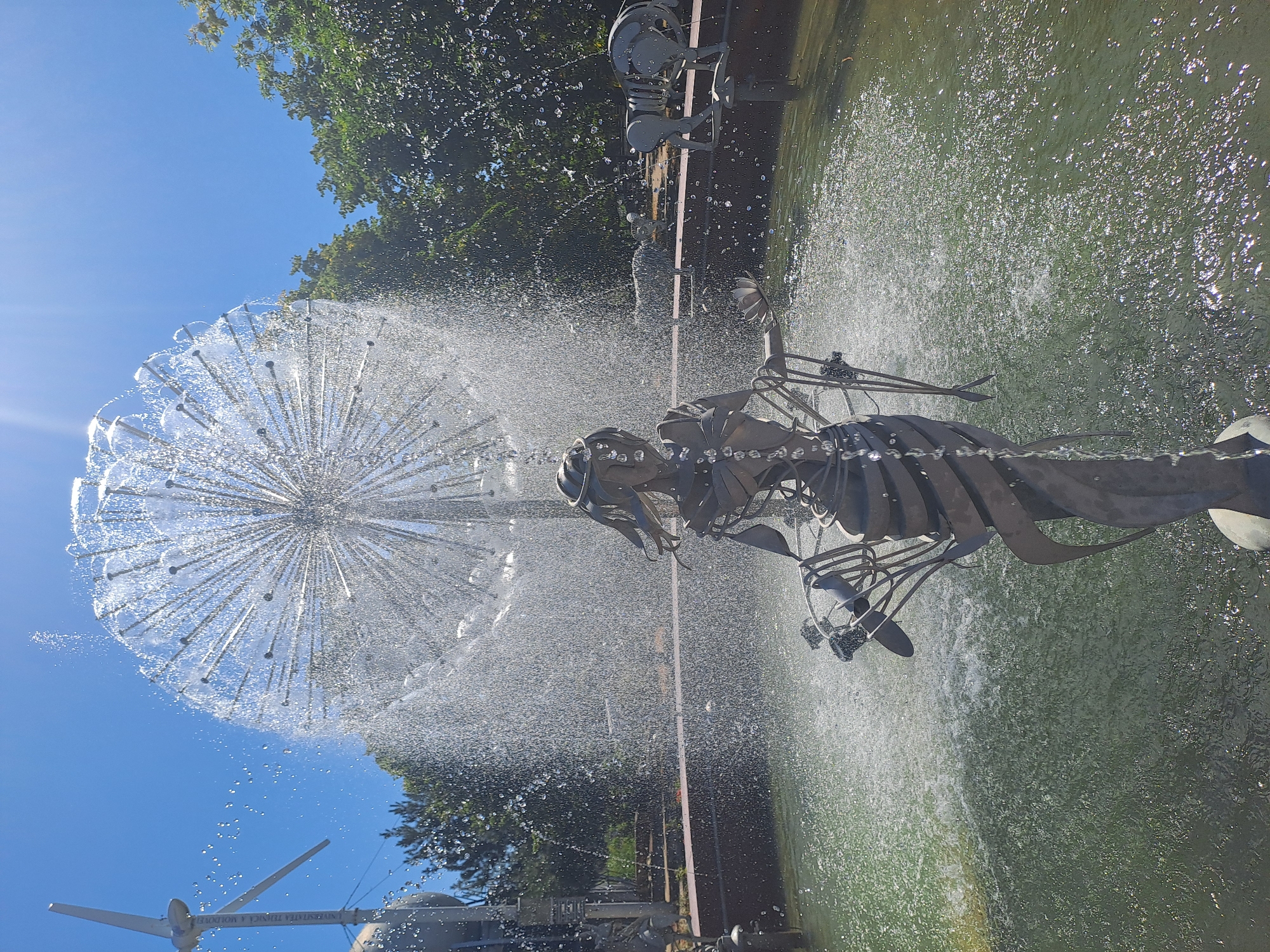 The image shows a fountain in Chișinău, Moldova, featuring a metal sculpture of a woman in the foreground and a large, spherical dandelion-shaped water feature behind her. The fountain is active, with water spraying in a wide arc from the spherical structure, creating a shimmering effect under the bright sunlight. The woman sculpture is stylized, with flowing lines that mimic fabric. She appears to be holding something in her hands, though the details are obscured by the spray. In the background, trees and other decorative elements of the park are visible. The water in the fountain pool is green and reflects the sunlight. A windmill-like structure is also visible to the left of the fountain.