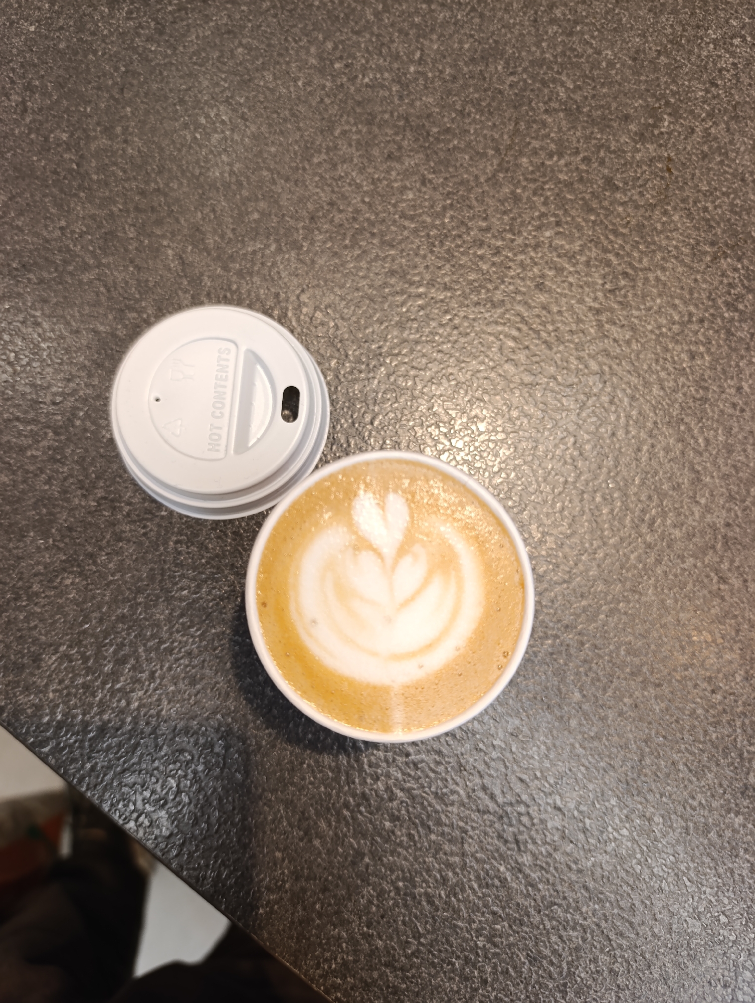 An overhead shot captures a single coffee cup with latte art and a white disposable lid on a dark, textured surface. The coffee, likely a latte, displays a well-executed heart or tulip design in the foam. The white lid, placed next to the cup, features embossed text reading "HOT CONTENTS" and has a small opening for drinking, as well as a smaller vent hole. The surface beneath the cup and lid appears to be a dark gray or brownish-gray material, possibly a countertop or table, with a slightly rough, granular texture. The lighting suggests an indoor setting, possibly a cafe. No people are visible, and no specific activities beyond the implied consumption of the coffee are depicted. The time of day and weather are not discernible. The image itself provides no visual cues to confirm the location in Sofia, Bulgaria.