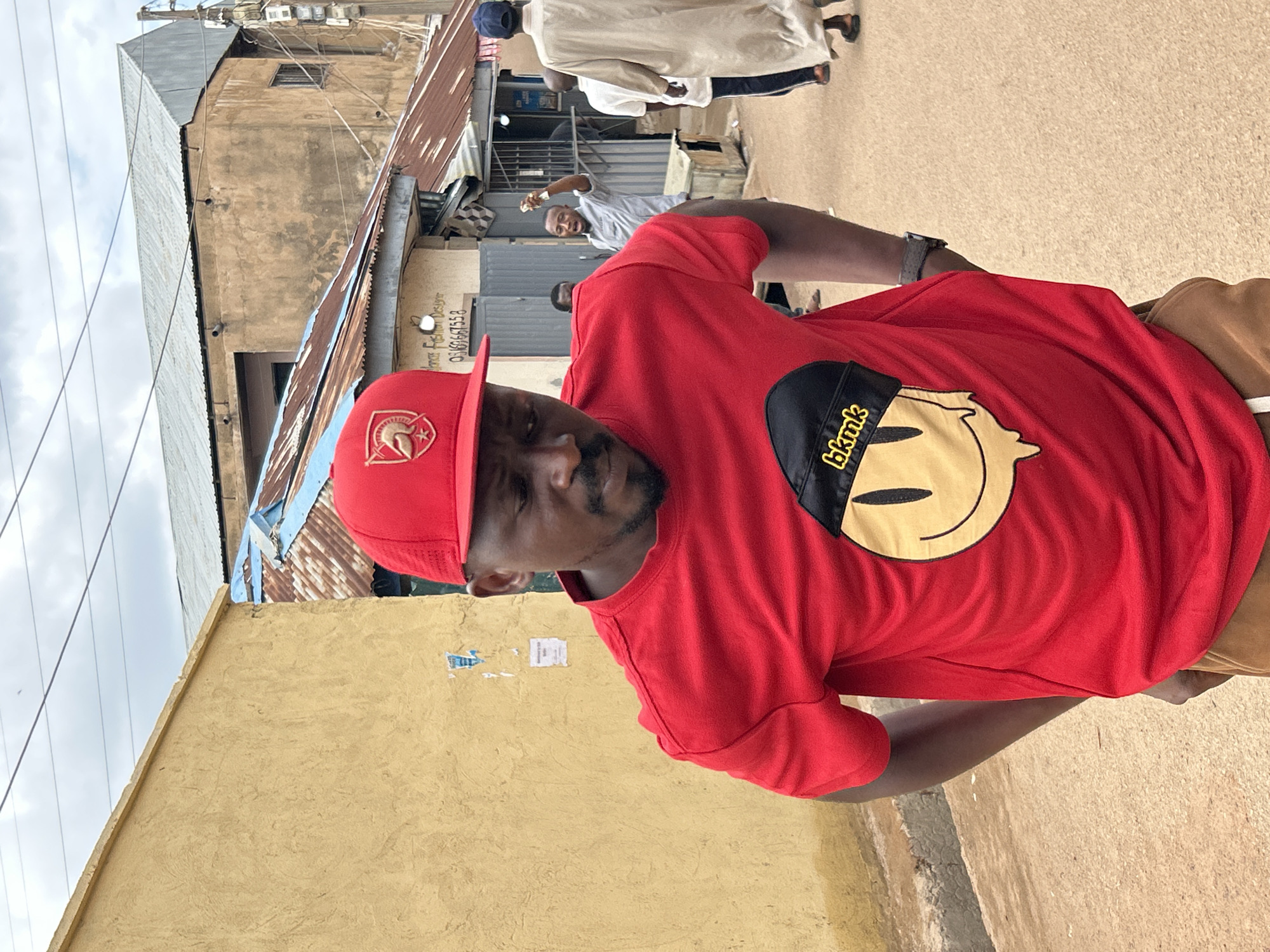 The image captures an urban street scene in Jos, Nigeria, under an overcast sky. In the foreground, a dark-skinned man is positioned slightly to the left of center, facing downwards and towards his right. He is wearing a red baseball cap adorned with a gold emblem, appearing to be a Spartan helmet, on the front. His attire includes a bright red short-sleeved t-shirt featuring a large graphic of a yellow smiley face wearing a black beanie with "bkmk" in yellow text, and the smiley face appears to be dripping at the bottom. Brown trousers are visible at his waist, and a grey-strapped watch is on his left wrist.
To the left of the central figure, a light yellow, textured building wall is visible, with remnants of torn posters or papers affixed to it. The background reveals a narrow, unpaved street or path. Several individuals are present in the mid-ground: a man in a light grey shirt is seen with his right arm raised, holding a small object, and his mouth open. Further to the right, a man in a long, light beige traditional robe (tobe or jalabiya) and a dark cap walks away from the viewer. Another person in a white t-shirt and dark pants is partially visible behind the robed man.
Buildings line the street; one low-rise structure features a rusted, corrugated metal roof with blue sections. A sign on its facade reads "Dance Fashion Designer" and displays the phone number "09169667558." Behind this, a larger, multi-story building constructed of weathered concrete with a galvanized corrugated iron roof is visible. A utility pole supporting multiple overhead wires stands on the far right side of the image. The ground is an unpaved, light brown surface.