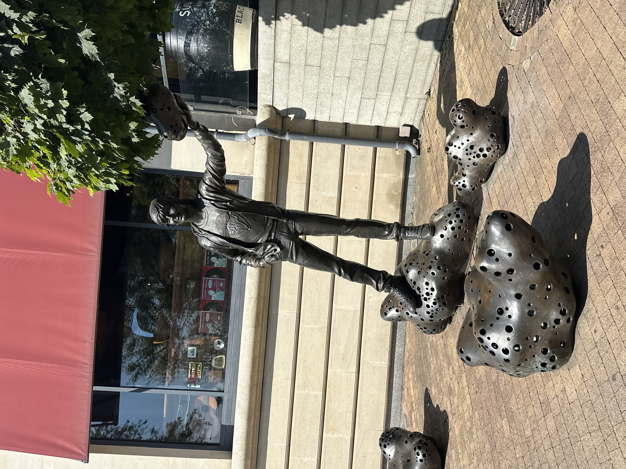 The image shows a bronze sculpture of a young man in a leather jacket and jeans, standing on a series of perforated, amorphous shapes. He is holding one of these perforated shapes above his head. The sculpture is located outdoors on a brick-paved area. In the background, there is a building with a red awning over a window display, and a tree with green leaves. A downspout is visible on the side of the building. The scene is brightly lit by sunlight, casting shadows on the ground. This sculpture is located in the city of Sochi, Russia.