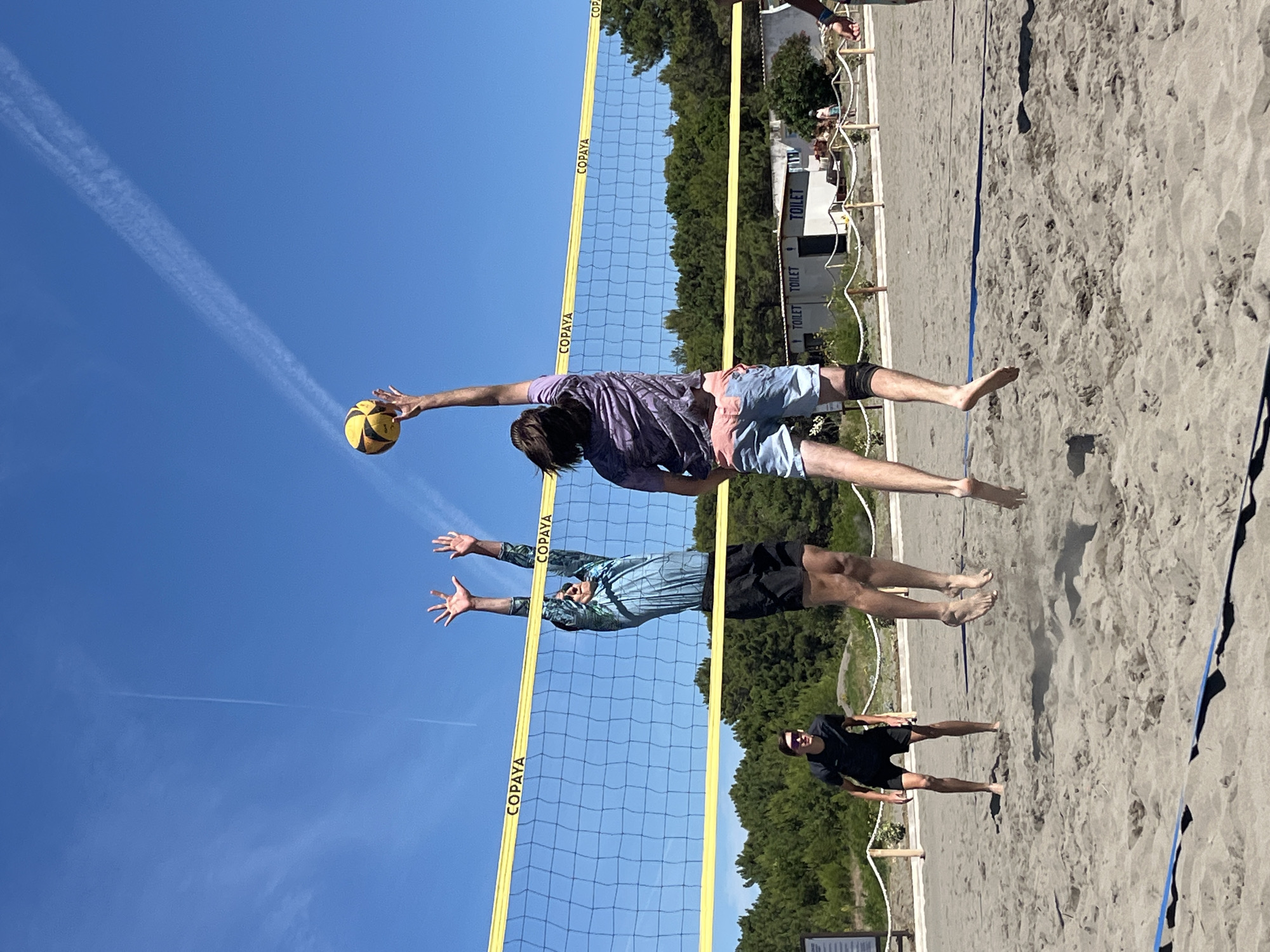 In Pistula, Montenegro, on a sunny day with a clear blue sky and a few wispy clouds, four individuals are engaged in a game of beach volleyball on a sand court. Two players are in mid-air, reaching for a yellow and black volleyball over a yellow-taped net. The player on the right, wearing a purple patterned shirt and light blue shorts with a knee brace, is jumping to spike the ball. The player on the left, in a blue patterned shirt and black shorts, is jumping to block. A third player, wearing a black t-shirt and black shorts, stands on the court to the left, facing the net, with sunglasses on. A fourth player, partially visible on the right, is also in motion. In the background, behind the court, a line of trees forms a dense green backdrop. Beyond the trees, there are a few buildings, one of which has signs that read "TOILET". The sand court has visible markings, including a blue line at the bottom of the frame.