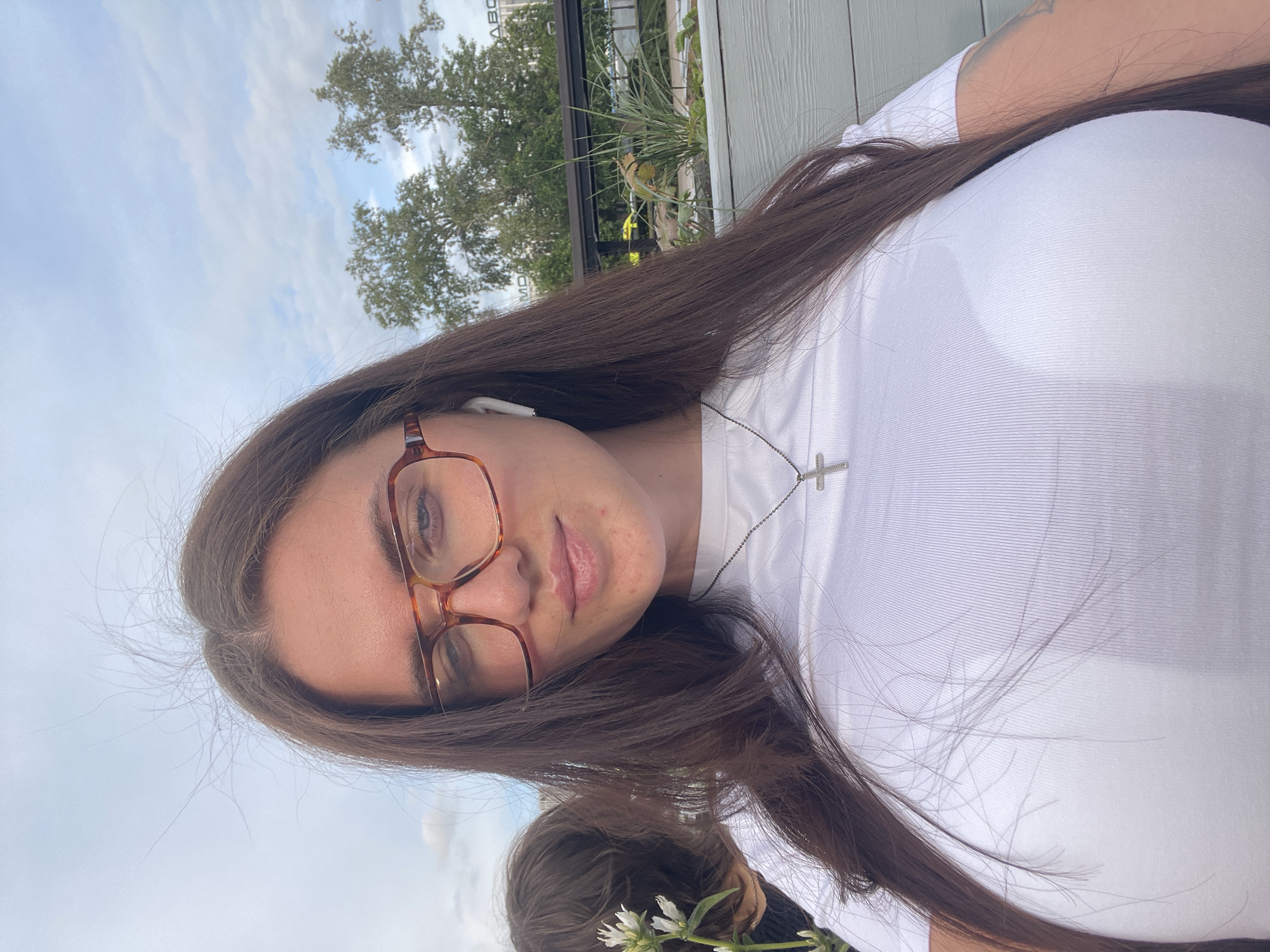 A person wearing glasses and a white shirt stands outdoors. The background includes trees, foliage, and a building. They are also wearing a cross necklace. The sky is visible with some clouds. The location is not identifiable as Moscow, Russia, from the given image.