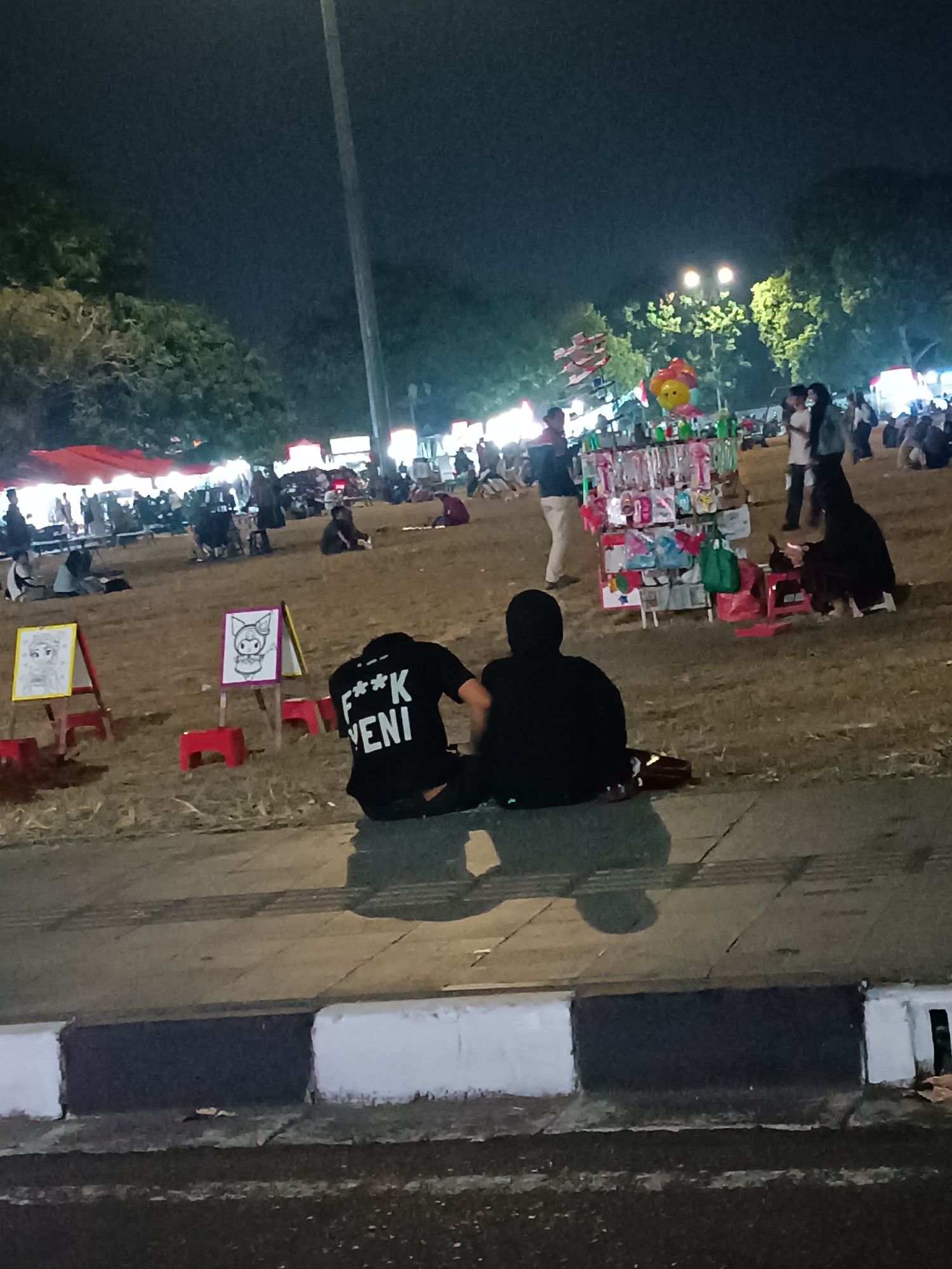 In Yogyakarta, Indonesia, at night, two individuals are seated on the ground in the foreground, their backs to the camera. The person on the left wears a black t-shirt with white text that reads "F*** VENI". The person on the right wears a black hooded garment, obscuring their face. Behind them, a variety of stalls are set up, some with red tarps providing shade. Several other people are visible in the middle ground, some browsing at the stalls and others sitting or walking. Further in the background, trees are visible, and lights from the stalls and potentially the city illuminate the scene. The ground is covered in dry grass and dirt. In the very front of the image, a concrete curb and pavement are visible.
