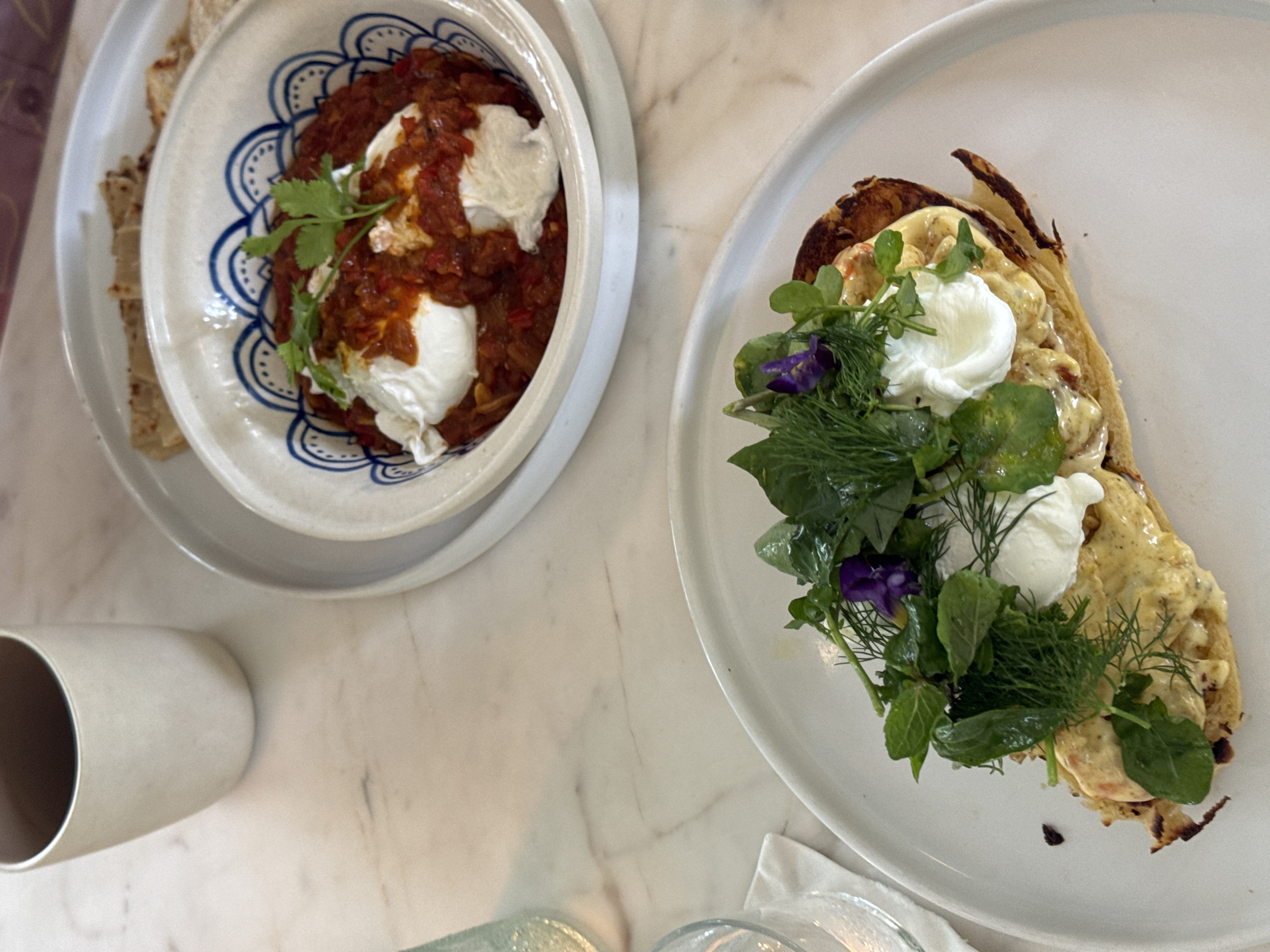 On a light marble surface, two dishes are presented. In the foreground, a white plate holds a dish topped with a creamy sauce, two poached eggs, and a garnish of fresh green herbs and small purple flowers. To the left of this plate, a part of a clear glass is visible. In the background, a white bowl with blue detailing contains a red sauce with visible pieces of vegetables and two more poached eggs, garnished with a sprig of cilantro. Beside this bowl, a stack of flatbreads is partially visible. A tall, light-colored mug sits to the left and slightly behind the background dish. The lighting suggests an indoor setting with soft ambient light. The overall scene depicts a breakfast or brunch setting. There are no people visible in the image. The location is identified as Kathaluwa, Sri Lanka.