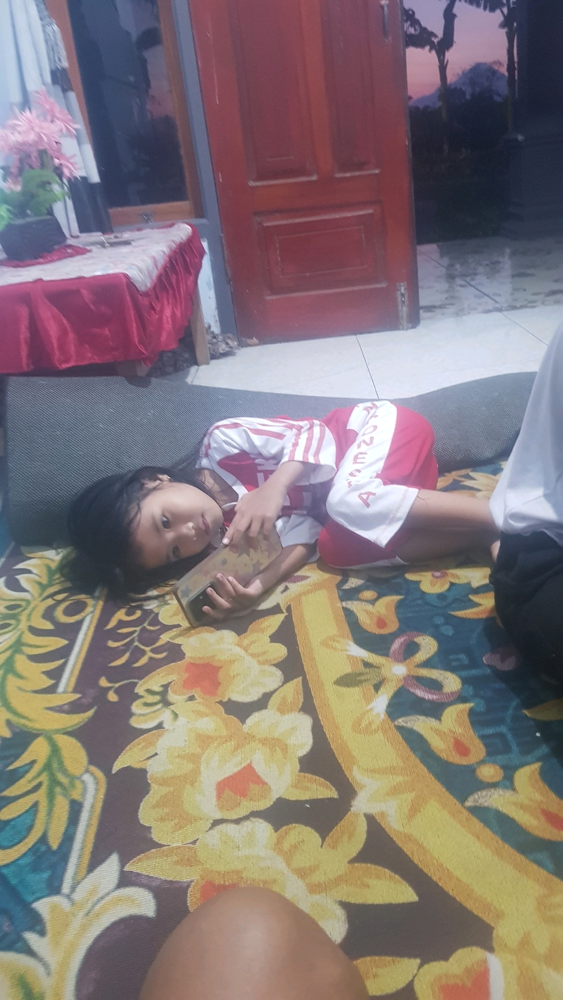 In Bakalan, Indonesia, a young girl lies on her side on a patterned rug, holding a phone in her hands. She wears a red and white striped shirt and red shorts with white lettering. The girl is positioned on what appears to be a thick, dark cushion or mattress. The background shows a red wooden door, part of a window with white curtains, and a table covered with a red cloth. Beyond the doorway, a dimly lit outdoor scene with silhouetted trees and a mountain is visible against a twilight sky. The overall impression is one of a casual, indoor setting with hints of the outdoors.