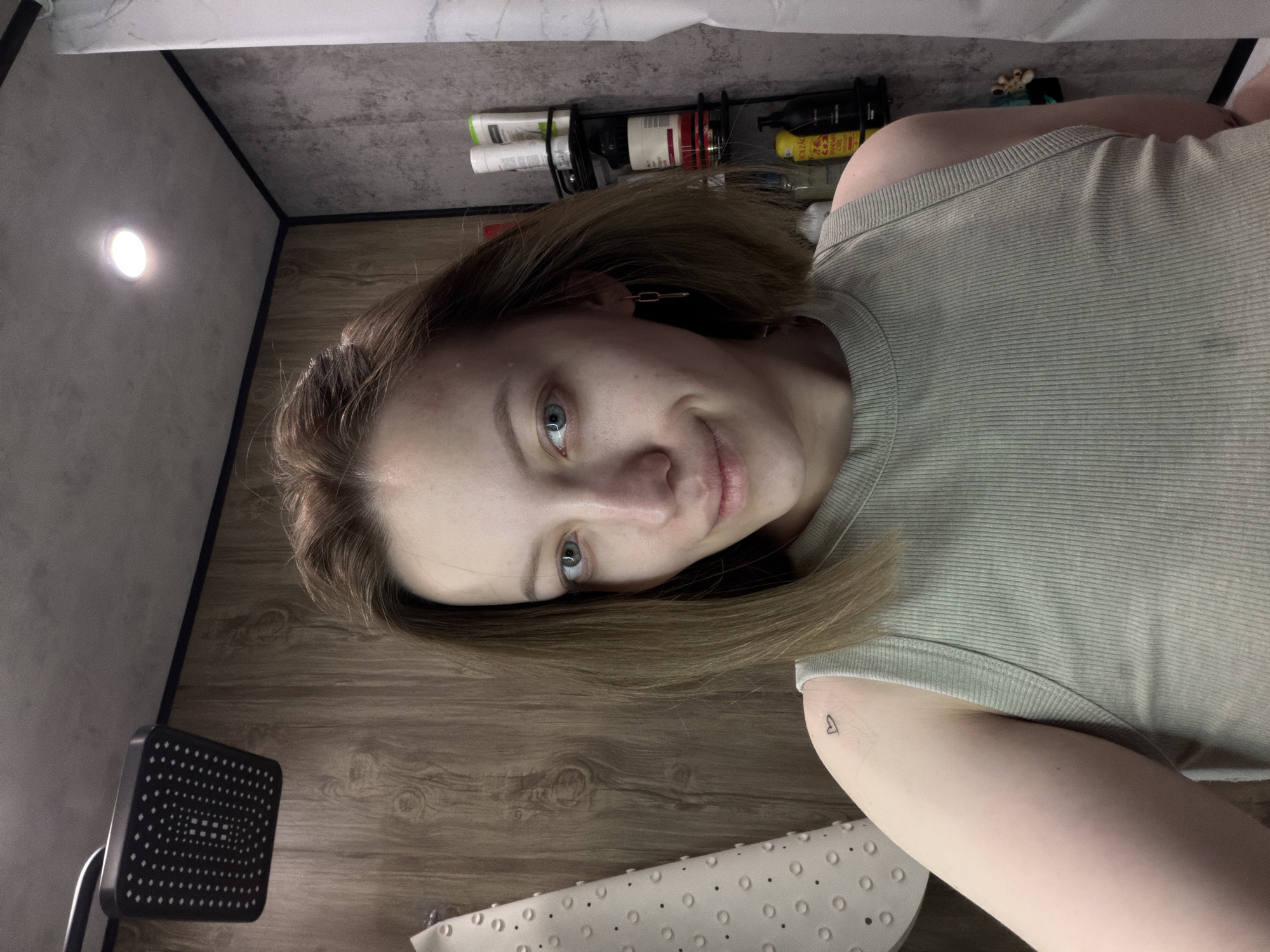 A woman is pictured in a bathroom. The bathroom has gray and wood-patterned walls. A showerhead is on the left. A shelf holding various bottles is visible in the background. The woman has shoulder-length brown hair and is wearing a light green top. A small heart tattoo is visible on her arm. The image is rotated. There is no indication of the location being Reshetnikovo, Russia.