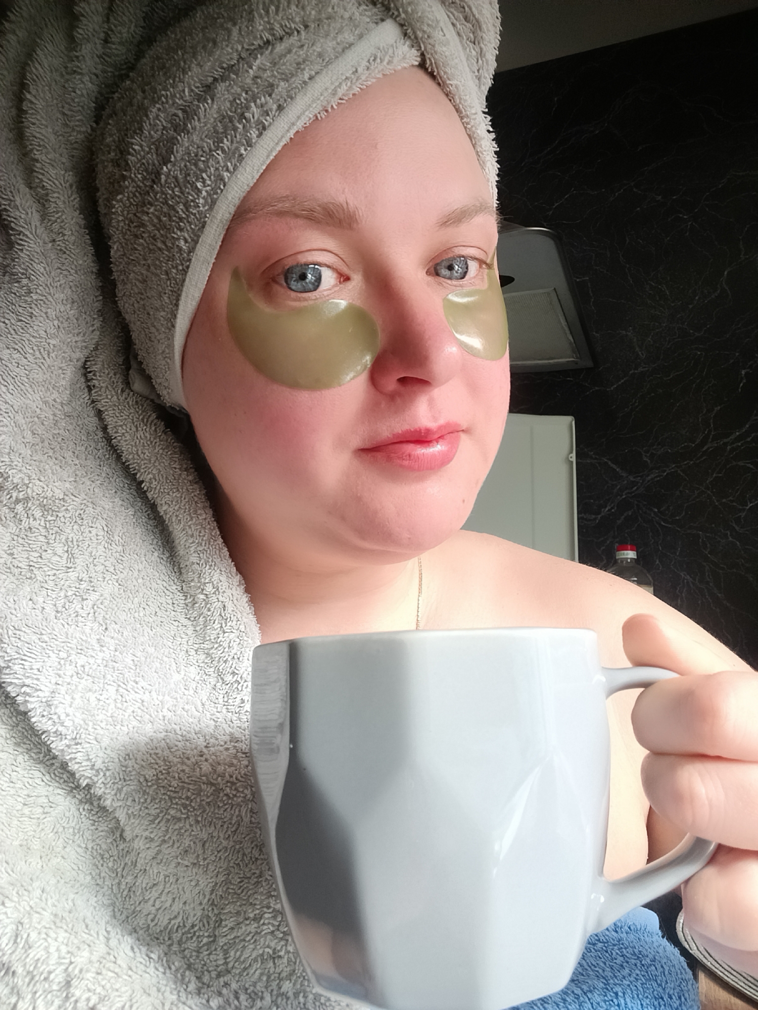 A fair-skinned woman with blue eyes is holding a gray mug and wearing green under-eye patches. She has a gray towel wrapped around her head. The background includes a black marble-patterned wall, a white cabinet, and a bottle with a red cap. The location is Chaikovsky, Russia.