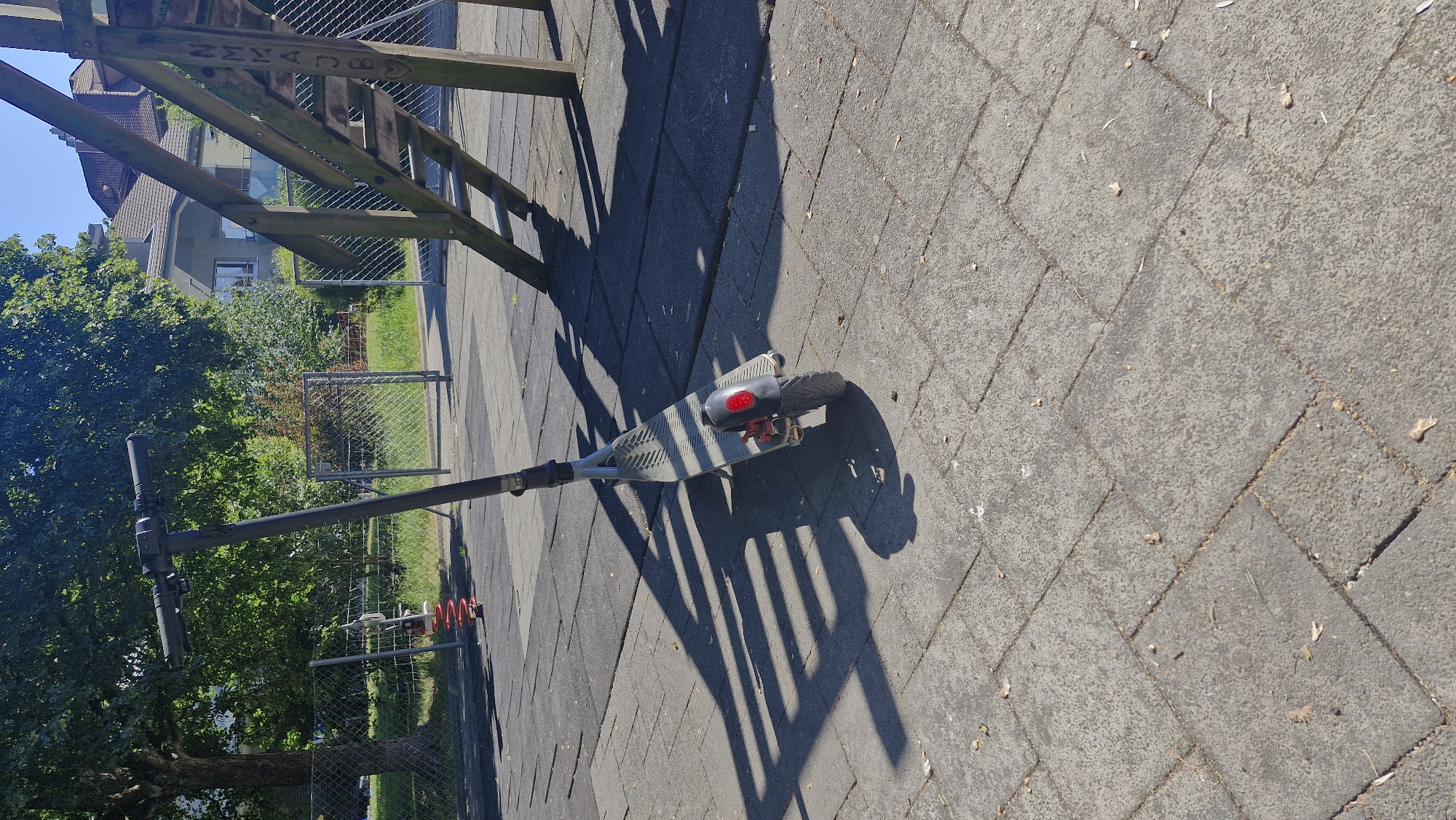 A dark gray electric scooter with a red taillight stands on a paved area in what appears to be a public park or playground. The scooter is positioned at an angle, with its shadow cast across the pavement, indicating a sunny day. Behind the scooter, a wooden playground structure with stairs is visible, and further in the background, a chain-link fence, trees, and buildings can be seen. The ground is paved with square concrete tiles. This scene is located in Wattenwil, Switzerland.