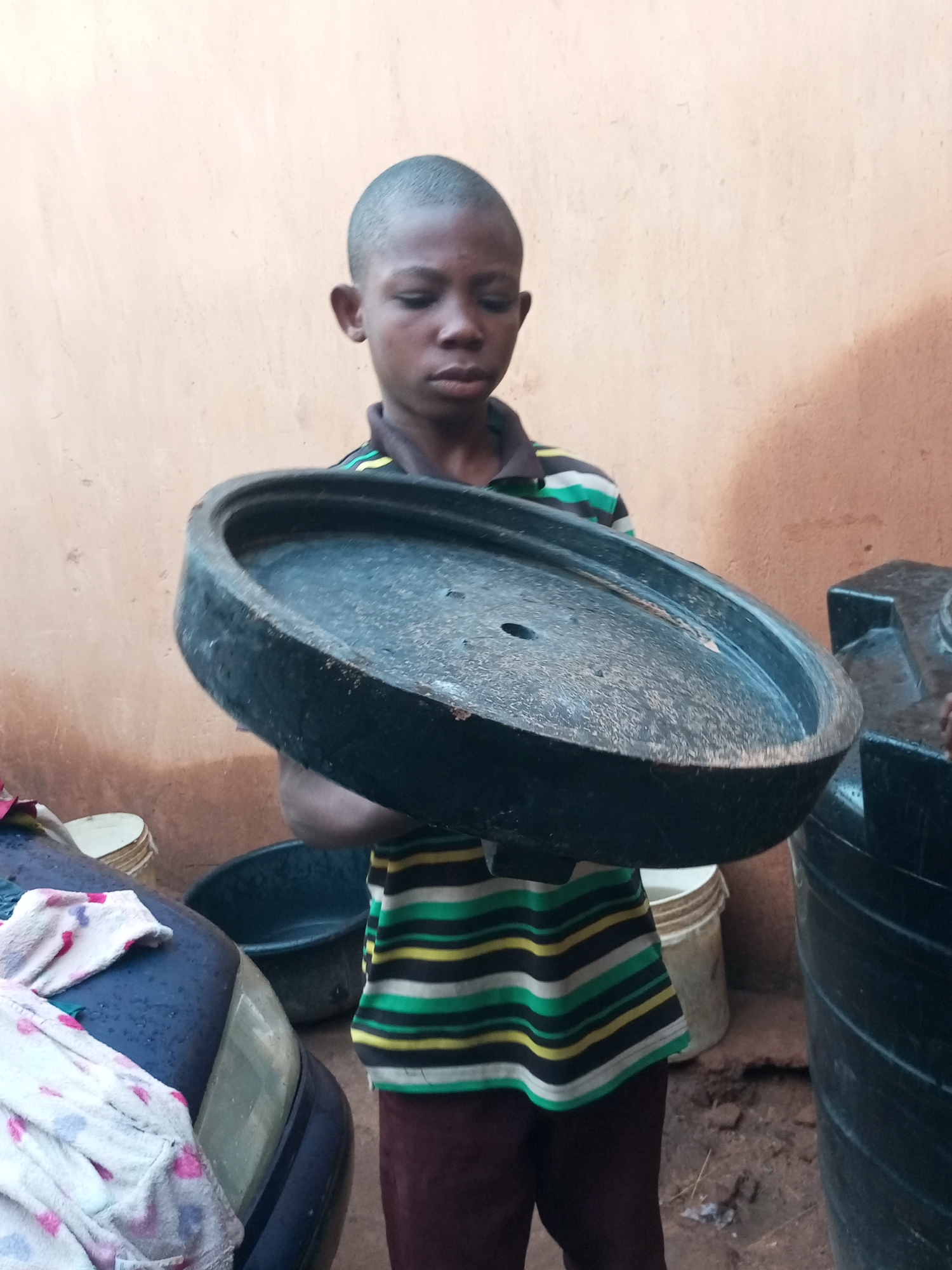 The image shows a young boy in Tempure, Nigeria, holding a large, circular, black lid with a hole in the center. He is wearing a striped t-shirt with green, yellow, and black bands, and dark brown pants. The boy appears to be standing outdoors, with a textured, light brown wall behind him. To his left, a portion of a dark blue car is visible, with a white towel draped over its hood. To his right, a large black water tank is present. Several buckets and containers are also scattered around the scene. The ground is uneven and appears to be made of dirt. The overall impression is one of a domestic or utilitarian outdoor space.