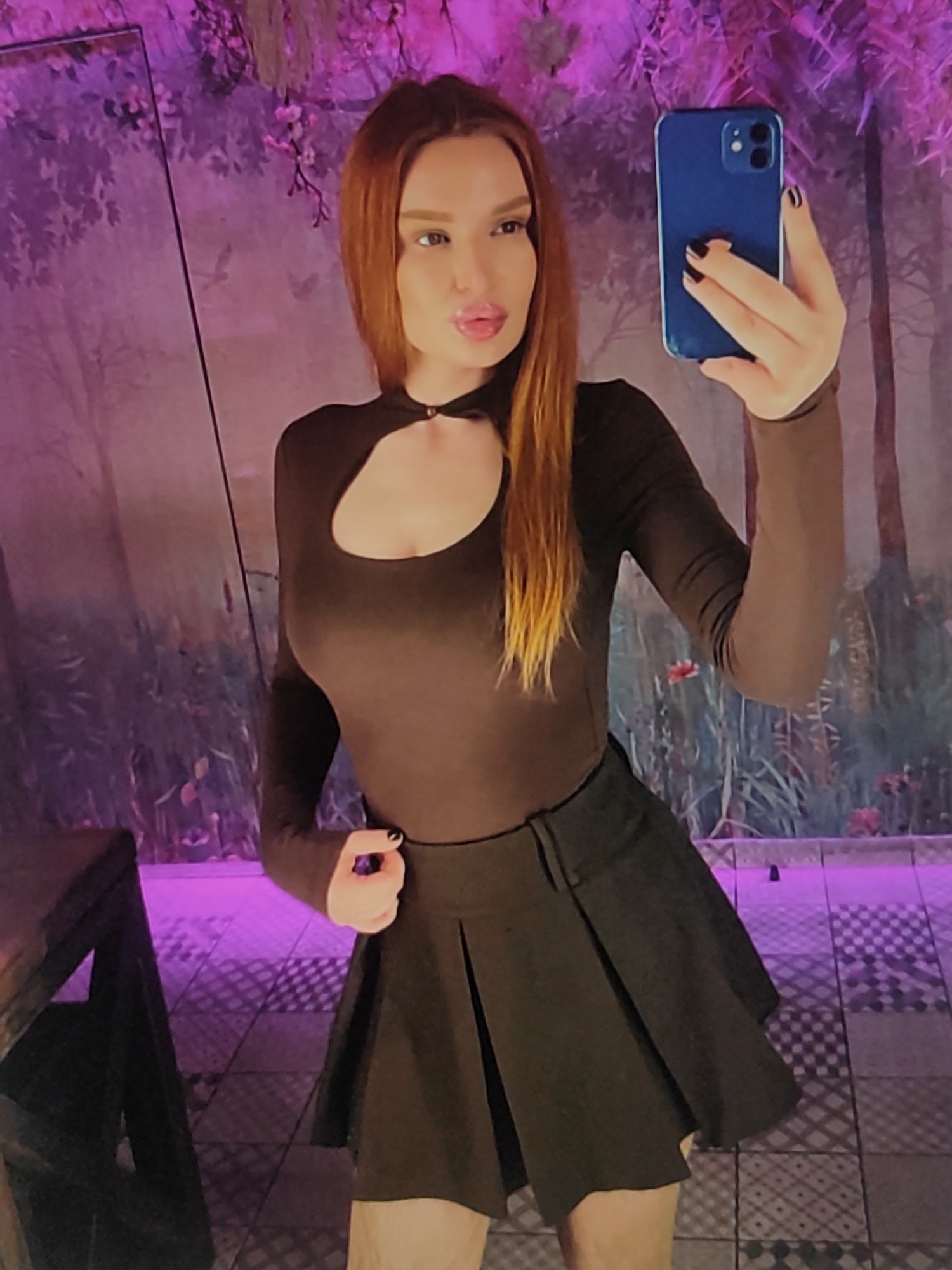 A young woman with auburn hair is taking a mirror selfie indoors, holding a blue smartphone in her right hand. She is wearing a brown, long-sleeved, form-fitting top with a cutout at the chest and a dark, pleated mini-skirt. Her fingernails are painted black.
The background features a wall with a mural depicting trees and flowers. The lighting is dim and predominantly purple, casting shadows on the floor, which is tiled in a geometric pattern. A dark wooden table is partially visible on the left side of the frame. The overall atmosphere suggests an evening setting, possibly in a dimly lit bar or lounge. There is no visible text in the image. The woman's expression is playful, with her lips pursed.
