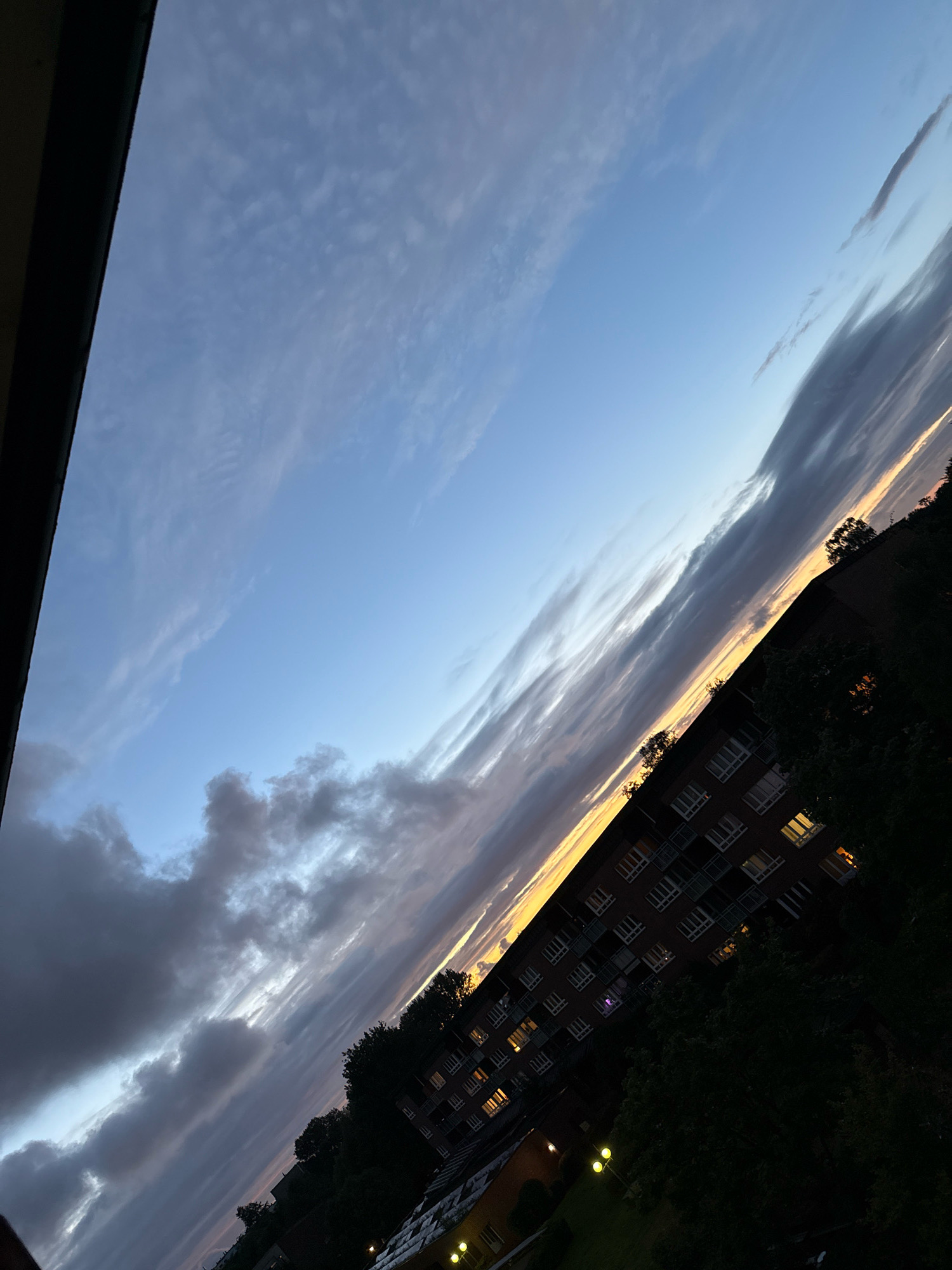 This is a diagonally oriented, low-angle photograph of a twilight sky over an apartment building in Hamburg, Germany. The sky is a gradient of blues and grays, with streaks of orange and yellow near the horizon, indicating a sunset. Wispy clouds are scattered across the sky. A multi-story apartment building with numerous lit windows is visible on the right side of the image, partially obscured by dark green trees. The building appears to be made of dark brick. In the foreground, on the left, a dark, solid object, possibly a balcony railing, is visible. The scene is peaceful and shows a typical urban evening view.