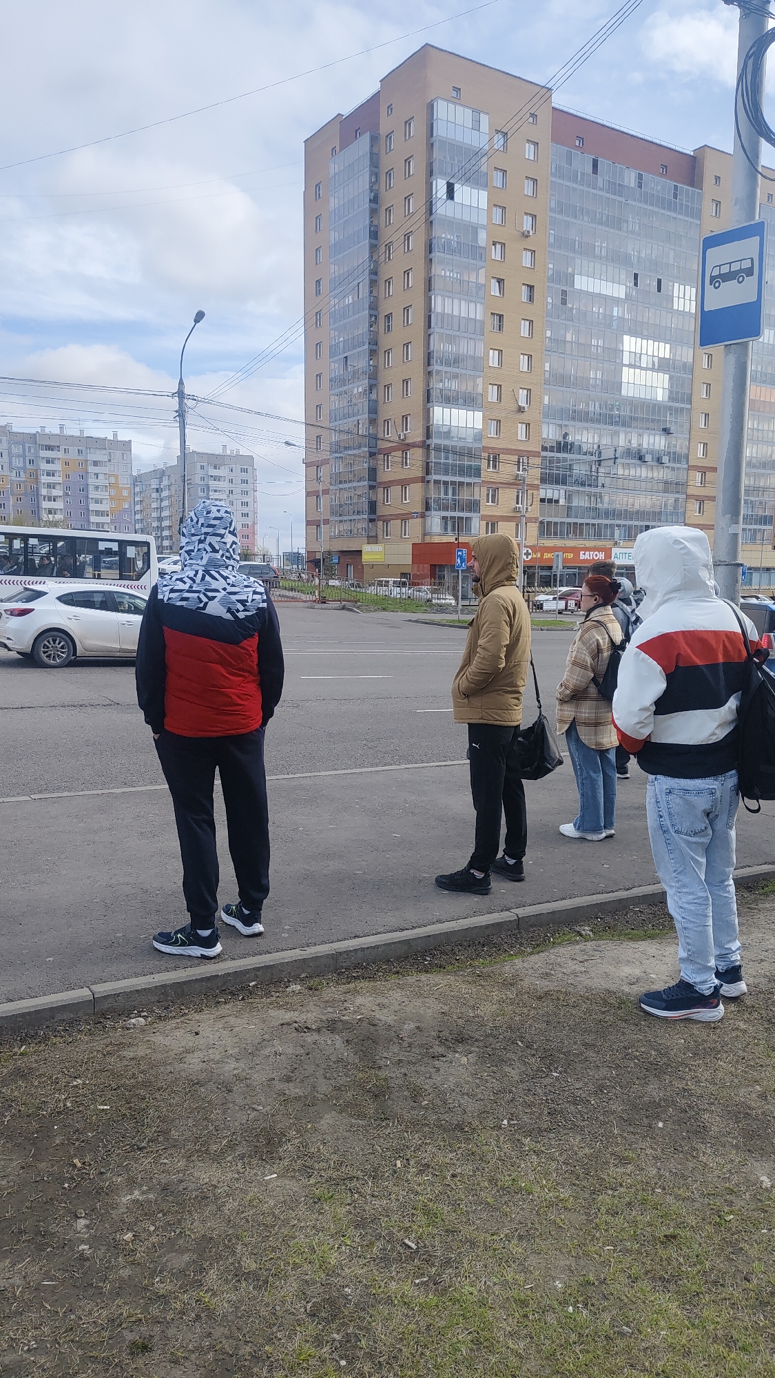 The image shows four people standing at a bus stop in Krasnoyarsk, Russia. They are waiting on a sidewalk next to a road. A tall, multi-story building with glass panels is in the background, along with other buildings in the distance. A bus is partially visible behind a white car. The bus stop sign is visible on a pole. The ground next to the sidewalk is a mix of dirt and sparse grass. The sky is overcast.
