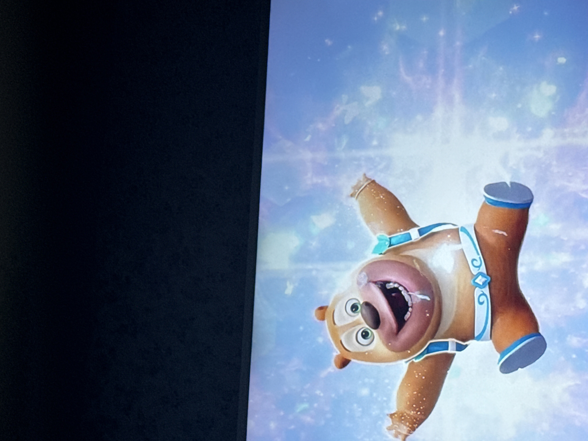 The image shows a cartoon bear character with its mouth open and arms and legs splayed, seemingly falling against a light blue background with a bright, abstract pattern. The bear is wearing what appears to be a blue and white harness with ornamental details. The image is displayed on a screen, with a dark, possibly black, object on the left side of the screen. The location is not discernible from the image and cannot be determined to be Mytishchi, Russia or anywhere else.