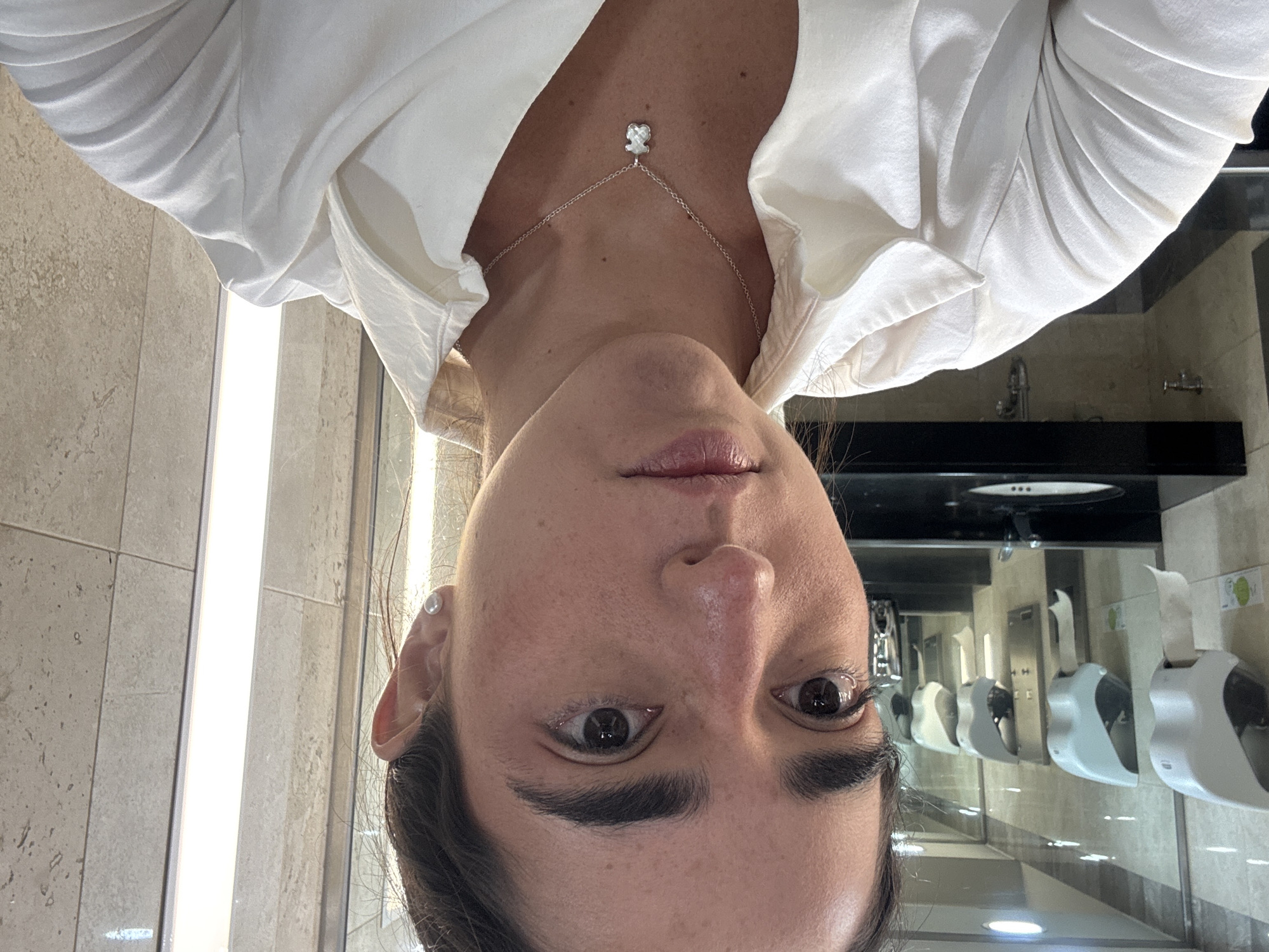 The image shows a selfie taken by a woman in what appears to be a public restroom. She is wearing a white blouse and a silver necklace with a small bear pendant. The background shows a row of automated paper towel dispensers mounted on a tiled wall, reflecting in a mirror above a black countertop with a sink. Given the restroom fixtures and tile design, and the prompt to include the location in Mexico City, Mexico, this is likely a restroom in a commercial or public establishment in that city.
