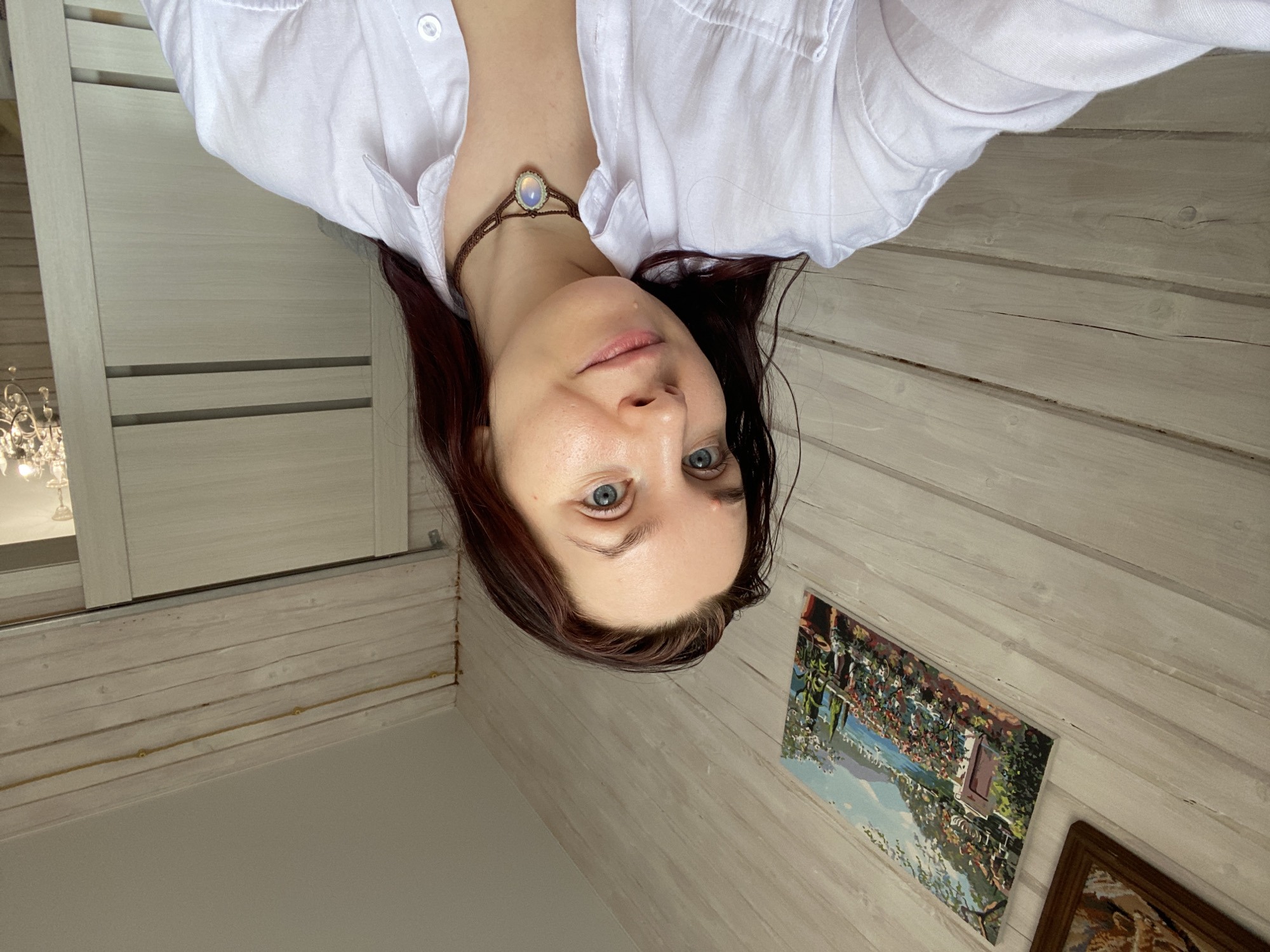 A woman with blue eyes and auburn hair wears a white button-down shirt and a brown necklace with a round pendant. She is indoors, posing for a selfie. The background is a whitewashed wood-paneled wall with two framed pictures. The location is possibly Фоки, Russia, based on the request, but not confirmed by the image itself. The scene appears to be in a room with a window and a chandelier visible in the upper left corner.