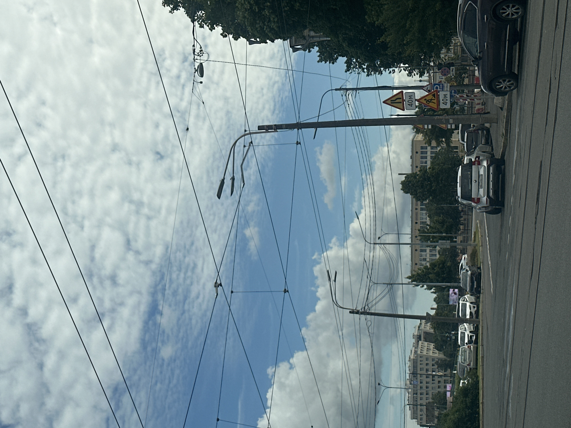 This is a street-level, eye-level, full-shot photograph taken in Saint Petersburg, Russia, on a cloudy day. The scene depicts a city intersection with a road in the foreground and a line of cars parked on the right side of the road. In the background, there are several buildings, trees, and a complex network of overhead wires, likely for public transportation.
**Specific details include:**
* **Vehicles:** Several cars are visible, including a dark-colored SUV on the far right, a white SUV, a silver SUV, and a darker sedan. Some of these cars are parked along the side of the road, while others appear to be in motion or waiting at the intersection.
* **Infrastructure:** Numerous overhead wires crisscross the frame, supported by poles. Streetlights are also visible, attached to some of these poles. Traffic signs are present, including signs indicating "40m" speed limit and road work.
* **Buildings:** In the background, there are multi-story buildings with windows, suggesting a commercial or residential area. Some of these buildings have architectural details consistent with Russian urban design.
* **Environment:** The sky is filled with white and gray clouds, with patches of blue sky visible. Trees line the sides of the road and are interspersed between buildings, indicating a green urban environment.
* **Road:** The road appears to be paved, with visible tram tracks running through it.
The overall impression is a typical urban scene in a Russian city, characterized by its infrastructure, vehicles, and architecture under a cloudy sky.