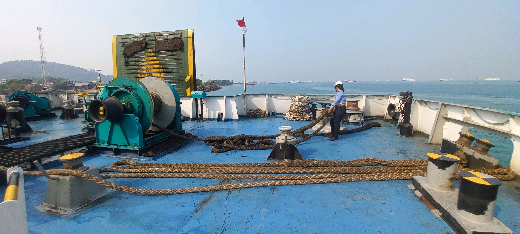 The image shows the deck of a cargo ship or ferry at sea, likely near Cilegon, Indonesia, given the distant view of a coastline with structures and a telecommunications tower. The deck is painted blue and is equipped with substantial ropes, winches, and bollards. In the foreground, thick, braided ropes are coiled and stretched across the deck. A person wearing a white hard hat, blue shirt, and dark pants is standing near the center of the frame, actively handling one of these thick ropes. To the left, a large, teal-colored winch with a coiled rope is prominent. In the background, the sea is visible with several other ships in the distance. A flag with red and white horizontal stripes, possibly the Indonesian flag, is flying on a pole. The sky is clear and bright. The overall scene depicts activity on a working ship.