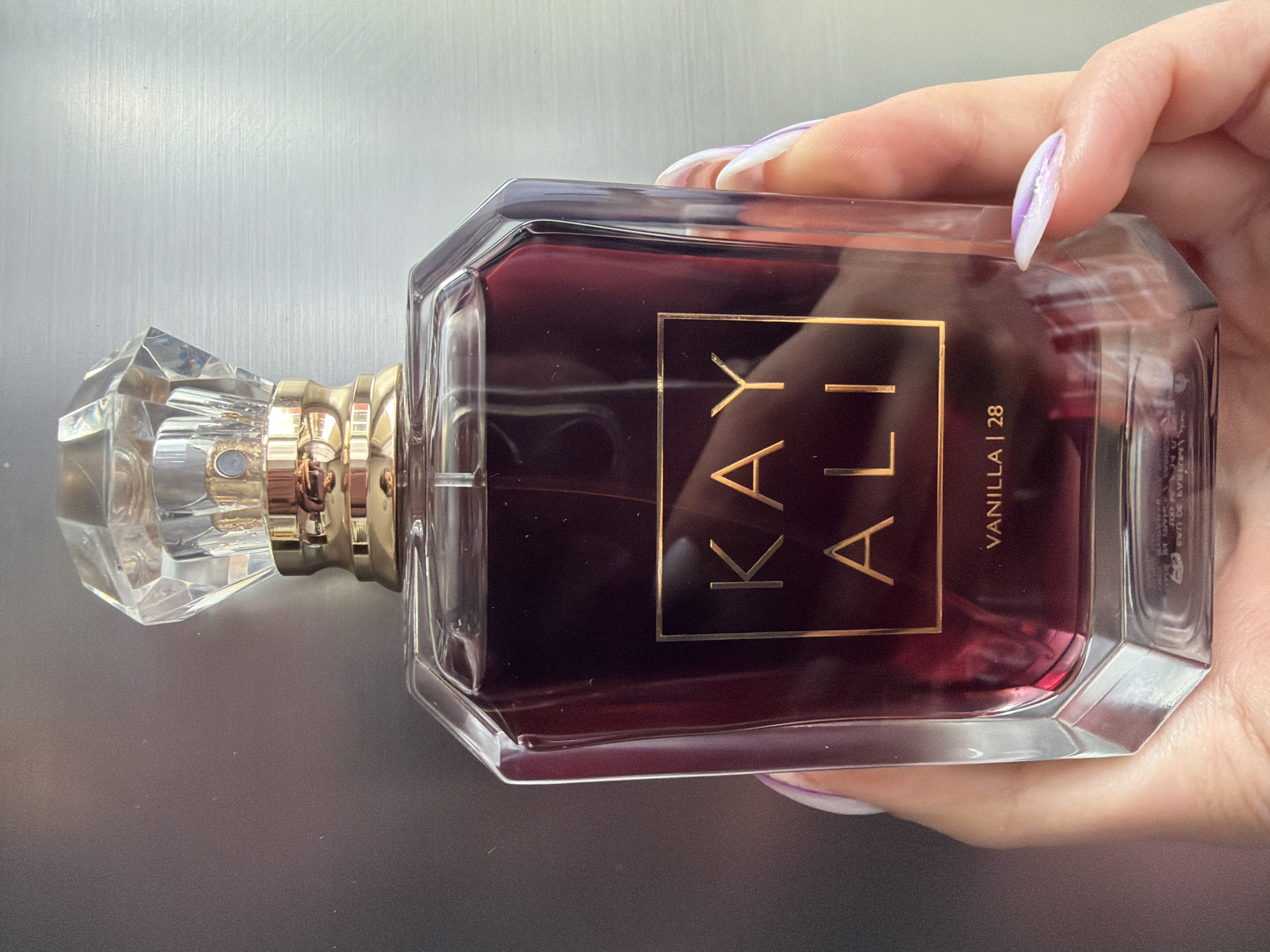 A hand is holding a bottle of Kayali Vanilla 28 perfume. The bottle is rectangular with a faceted crystal cap and gold accents. The liquid inside is a dark reddish-purple color. The background is a plain, light gray surface. The location is not discernable from the image and there is no indication it is in Pazardzhik, Bulgaria.