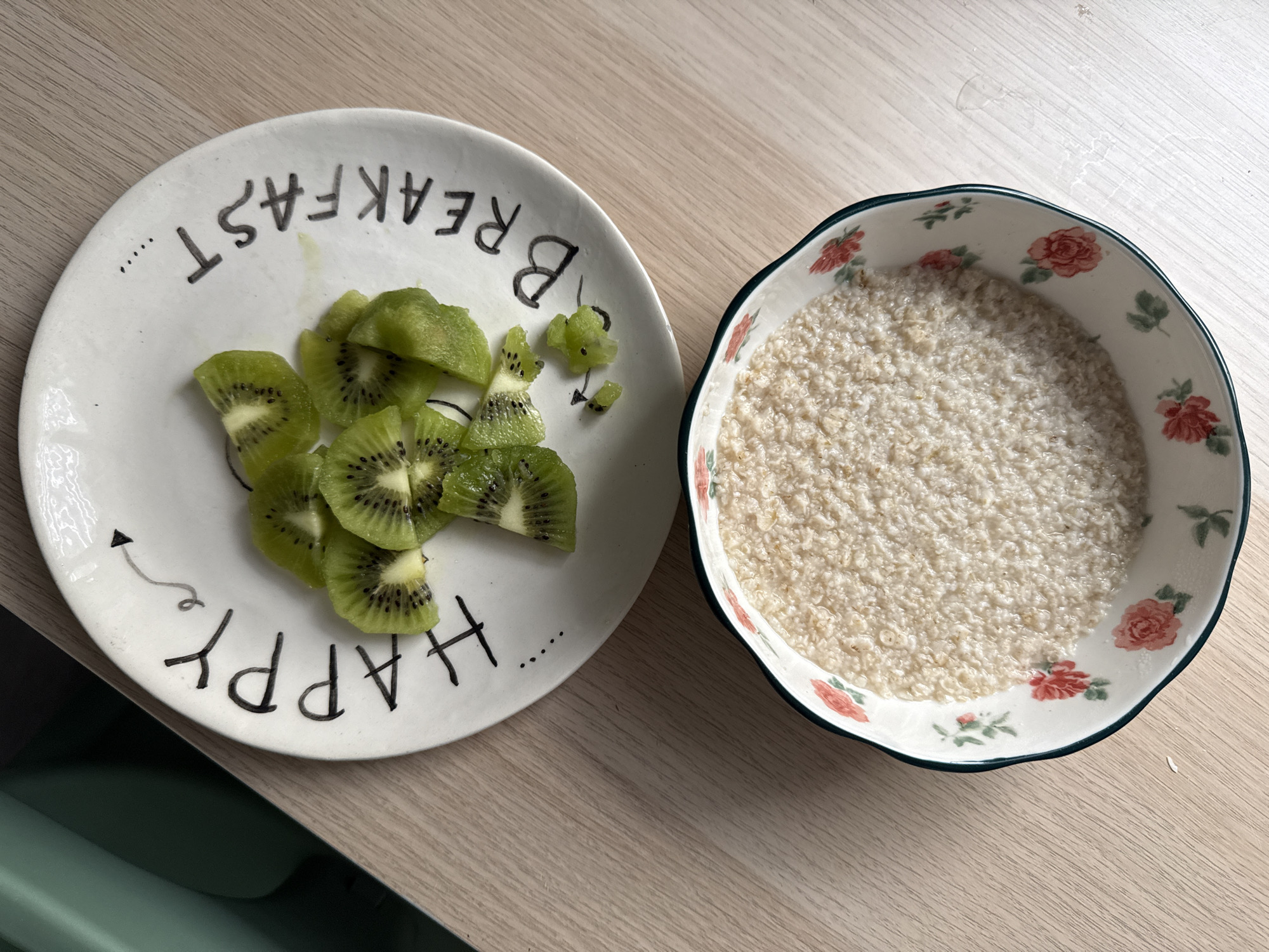 The image displays a simple breakfast or snack laid out on a light-colored wooden table. On the left, a round white ceramic plate holds several slices of fresh, green kiwi fruit. The kiwi slices are mostly whole, revealing their characteristic black seeds and light green flesh. The plate has handwritten-style black text around its rim, which, when fully read by rotating the plate, spells out "HAPPY BREAKFAST" along with an arrow and dots. Some text is visible right-side up ("HAPPY") and some inverted ("BREAKFAST"). On the right side of the image, there is a heart-shaped or scalloped ceramic bowl filled with a light-colored, granular food, likely oatmeal or porridge, given its texture. This bowl features a delicate floral pattern of red roses and green leaves around its interior rim. The lighting in the image suggests daytime, illuminating the food and tableware clearly on the neutral wooden surface. The scene is static, showcasing prepared food, likely just before consumption.