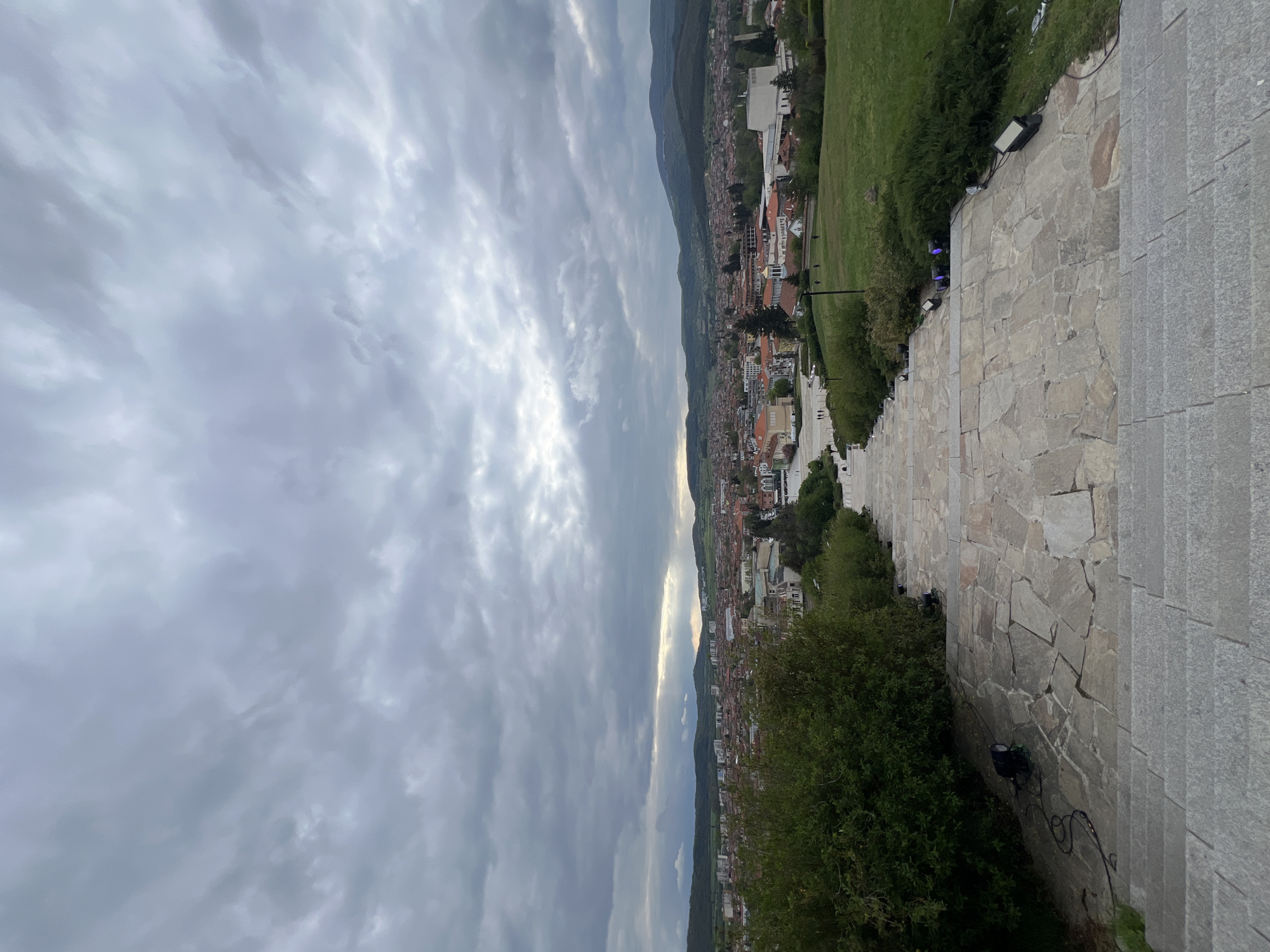 A panoramic view of the town of Panagyurishte, Bulgaria, is captured from a high vantage point under a cloudy sky. The scene is presented as if correctly oriented, though the original image is rotated.
In the foreground and to the right, a wide, gently sloping stone-paved path or staircase dominates. It is constructed from irregular, light-colored flagstones, bordered on its outer edge by more uniform grey stone, suggesting steps in some sections. Along the left side of this path, lush green grass slopes downwards, interspersed with dense green bushes and trees. Several outdoor floodlights are visible nestled among the foliage and beside the path, indicating nighttime illumination.
The midground reveals the sprawling town, characterized by a dense cluster of buildings, many featuring distinctive orange and red-tiled roofs. The urban area stretches horizontally across the valley, with some modern-looking white buildings interspersed among the more traditional structures.
In the background, rolling hills or mountains frame the valley, complementing the urban landscape. The sky above is heavily overcast with a thick layer of grey and white clouds. Patches of brighter light manage to pierce through the cloud cover in the distance, casting a soft, diffused glow over parts of the scenery and suggesting the sun's position.
The time of day appears to be daytime, possibly late afternoon, given the diffused light and cloud cover. The weather is calm and cloudy. A few indistinct figures of people can be seen walking on the stone path further in the distance. No discernible text is visible in the image. The overall impression is one of a serene landscape, blending urban development with natural surroundings.