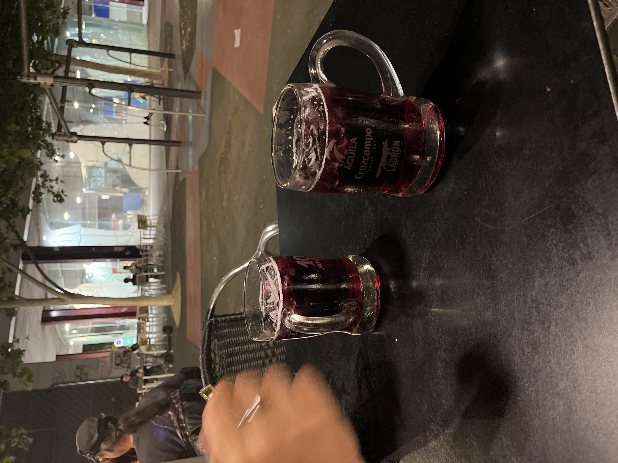 This image, captured from a slightly elevated perspective and rotated 90 degrees counter-clockwise, portrays an outdoor evening scene at a cafe or bar in Torrent, Spain. The primary subjects are two clear glass mugs with handles, filled with a dark red liquid, likely tinto de verano or red wine, and topped with some froth. They rest on a dark, possibly black or dark gray, table.
The closer mug prominently displays several brand names: "AGUILA", "CRUZCAMPO", "DESDE 1904", and "LADRÓN". The second mug, positioned further back, also shows "LADRÓN" partially. In the foreground, a hand wearing a golden ring is visible, slightly blurred due to motion, suggesting it is reaching for one of the drinks. A section of a black wicker chair is also visible on the left.
To the left, a person wearing a cap is partially visible, seated at the table. The background features an urban environment at night, with brightly lit interiors of a building seen through large windows. Trees with dark foliage and metallic structures, possibly outdoor lighting or part of an awning, are discernible against the illuminated backdrop. The ground appears paved. The overall scene suggests a relaxed social gathering, enjoying beverages outdoors during the evening.
