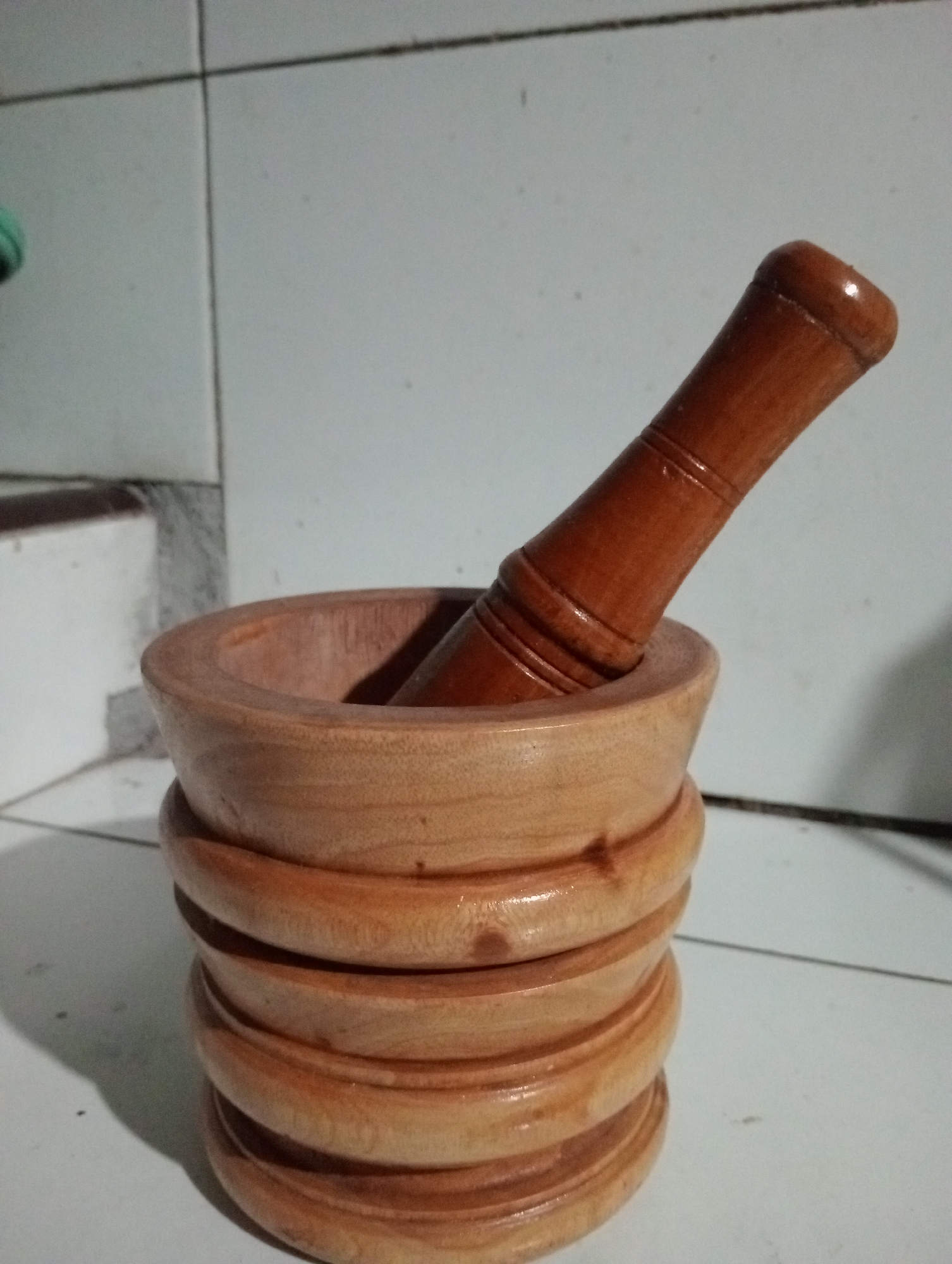 A wooden mortar and pestle are placed on a tiled surface. The mortar is cylindrical with stacked, rounded grooves along its exterior, and it contains the pestle. The pestle is also made of wood and has a rounded top and a grooved shaft. The background consists of light-colored square tiles with dark grout lines. The lighting creates shadows, suggesting an indoor setting. The image does not provide any information that would allow for its location to be determined in the city of Trenggalek, Indonesia.