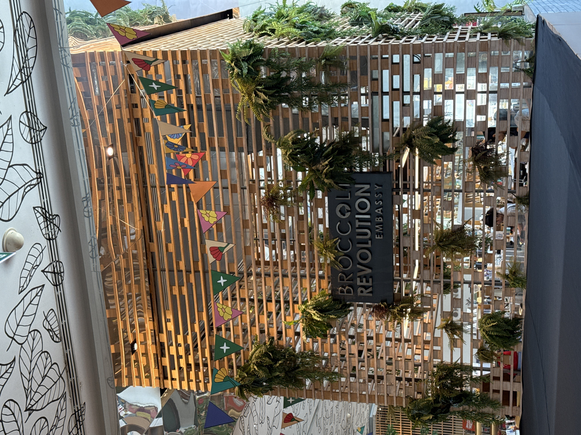 This is an interior architectural scene. A modern structure, possibly a store facade or an installation, is constructed with vertical wooden slats and a rectangular latticework pattern. The structure is decorated with green ferns and foliage. A dark sign on the structure reads "BROCCOLI REVOLUTION EMBASSY" vertically. Colorful triangular pennants hang along one side of the structure. The background reveals a blurred interior, reflecting figures and objects suggesting a commercial space. The scene is likely in Bangkok, Thailand, given the presence of the Broccoli Revolution restaurant and its branch location is in Bangkok, and the architectural style is congruent.