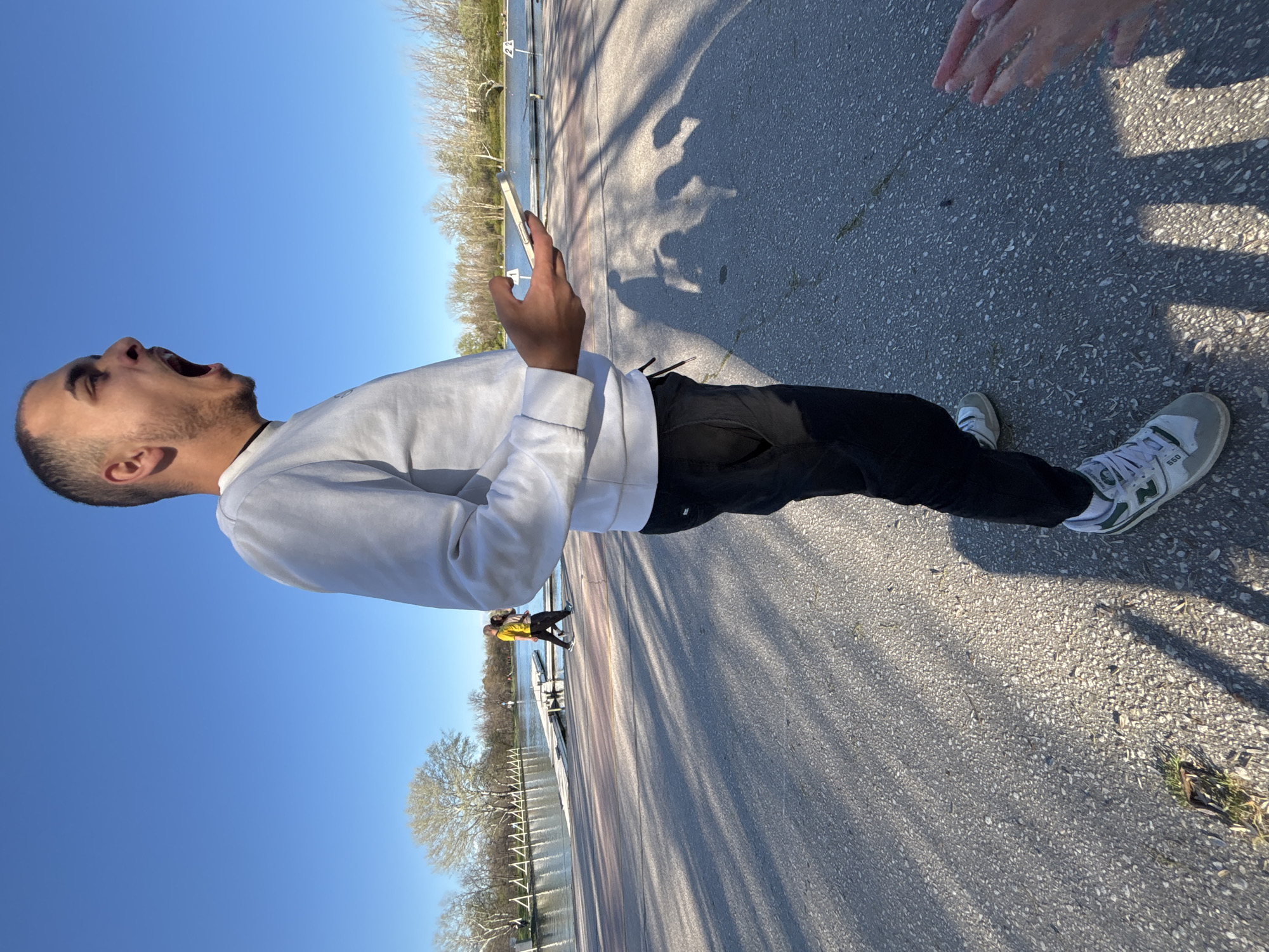 A man with a shaved head, wearing a white sweatshirt and black pants, stands on an asphalt surface near a body of water. He is holding a mobile phone and appears to be yelling. The sky is clear and blue. A couple can be seen walking along a path in the background. Another set of hands can be seen reaching towards the camera. There are trees and a fence along the water's edge. The location appears to be a park or recreational area. The specific location in Plovdiv, Bulgaria is unidentifiable from the image.