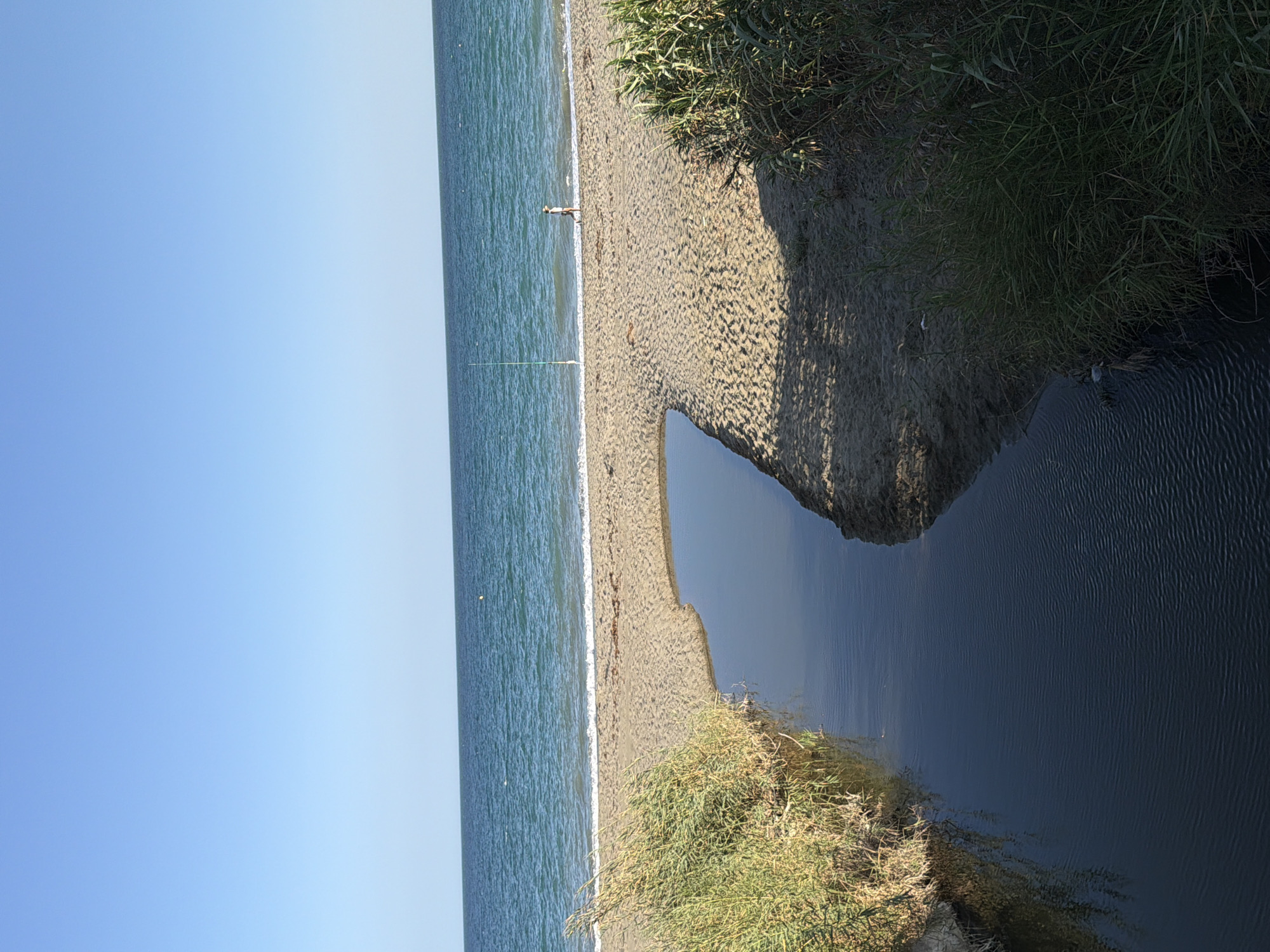 The image shows a wide, sandy beach with the Mediterranean Sea in the background. A small, dark stream flows into the sea, bordered by tall, dry grasses on the left and greener, denser vegetation on the right. The sand is mostly flat, with some footprints visible. In the distance, a single person wearing a hat and white top is walking along the shoreline, with a fishing rod standing upright nearby. The sky is clear and blue, with no clouds. The location is Saladillo-benamara, Spain.