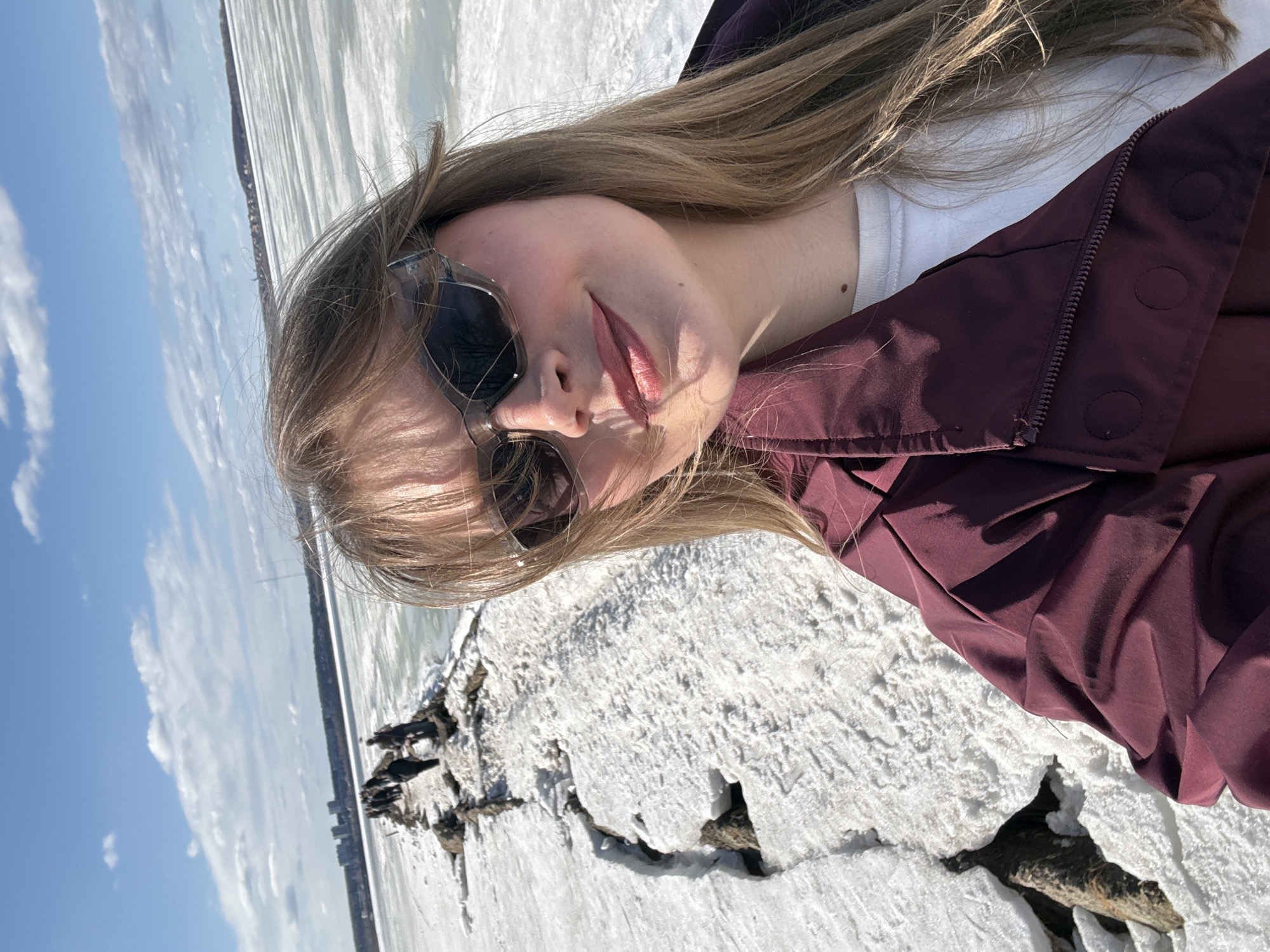 A woman with long blonde hair and sunglasses is taking a selfie outdoors. She is wearing a maroon jacket over a white shirt. She stands on a snow-covered surface, possibly ice, with visible cracks and dark areas beneath the snow. The background features a partially frozen body of water, possibly a river or lake. On the opposite shore, a cityscape is faintly visible under a blue sky with clouds. The location appears to be near the frozen body of water on the outskirts of Izhevsk, Russia.