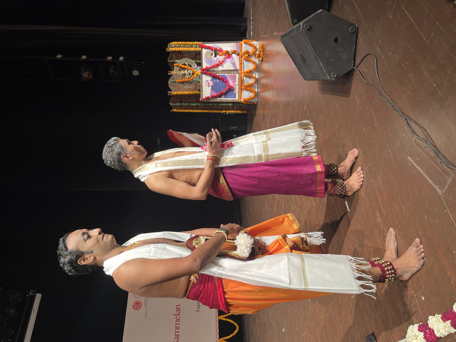 Two men stand on a stage in the Tagore Theatre, located in Sector 18, Chandigarh, India. They are both wearing traditional Indian clothing. The man on the left is holding a trophy. The man on the right is holding a piece of paper. There are two framed photographs behind them and several loudspeakers on the stage. The backdrop of the stage is a dark curtain. The floor is made of wood and the stage is brightly lit.