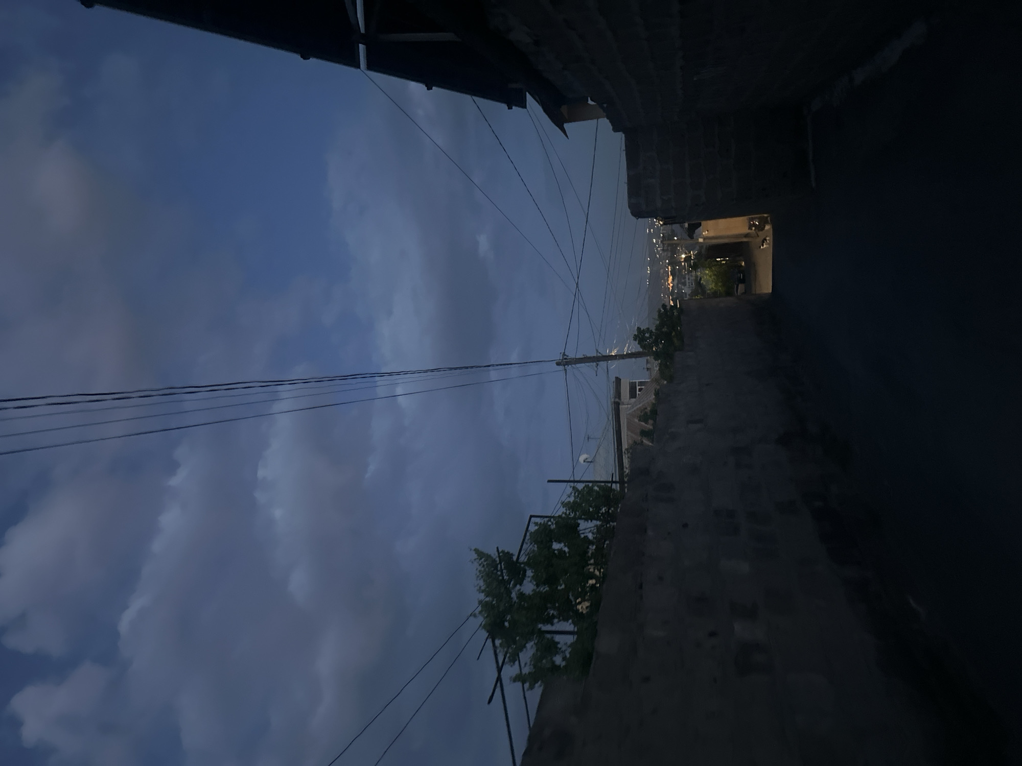 The photograph depicts a narrow alleyway in Yerevan, Armenia, at dusk. The sky, a gradient of blue and gray, is filled with clouds and crisscrossed by electrical wires. The alley is flanked by high stone walls, casting long shadows. At the end of the alley, a patch of light reveals a view of the city, with illuminated buildings visible in the distance. The edges of structures are partially visible at the top of the frame. In the middle of the frame, there is greenery on a trellis along the alleyway, and a pole is visible behind it.