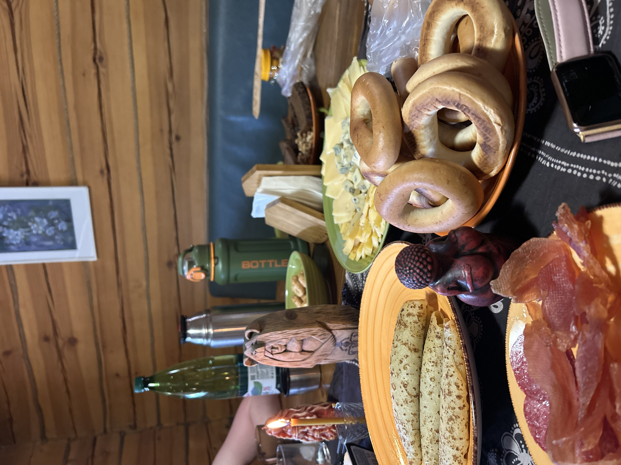 The image captures a table setting with a variety of food items, suggesting a meal or gathering. The food includes bagels, cheeses, pancakes, and sliced fish. A wooden carving and a candle are also present on the table. A thermos, a bottle of olive oil, and a framed picture are visible in the background. The location is Лесные Поляны, Russia.