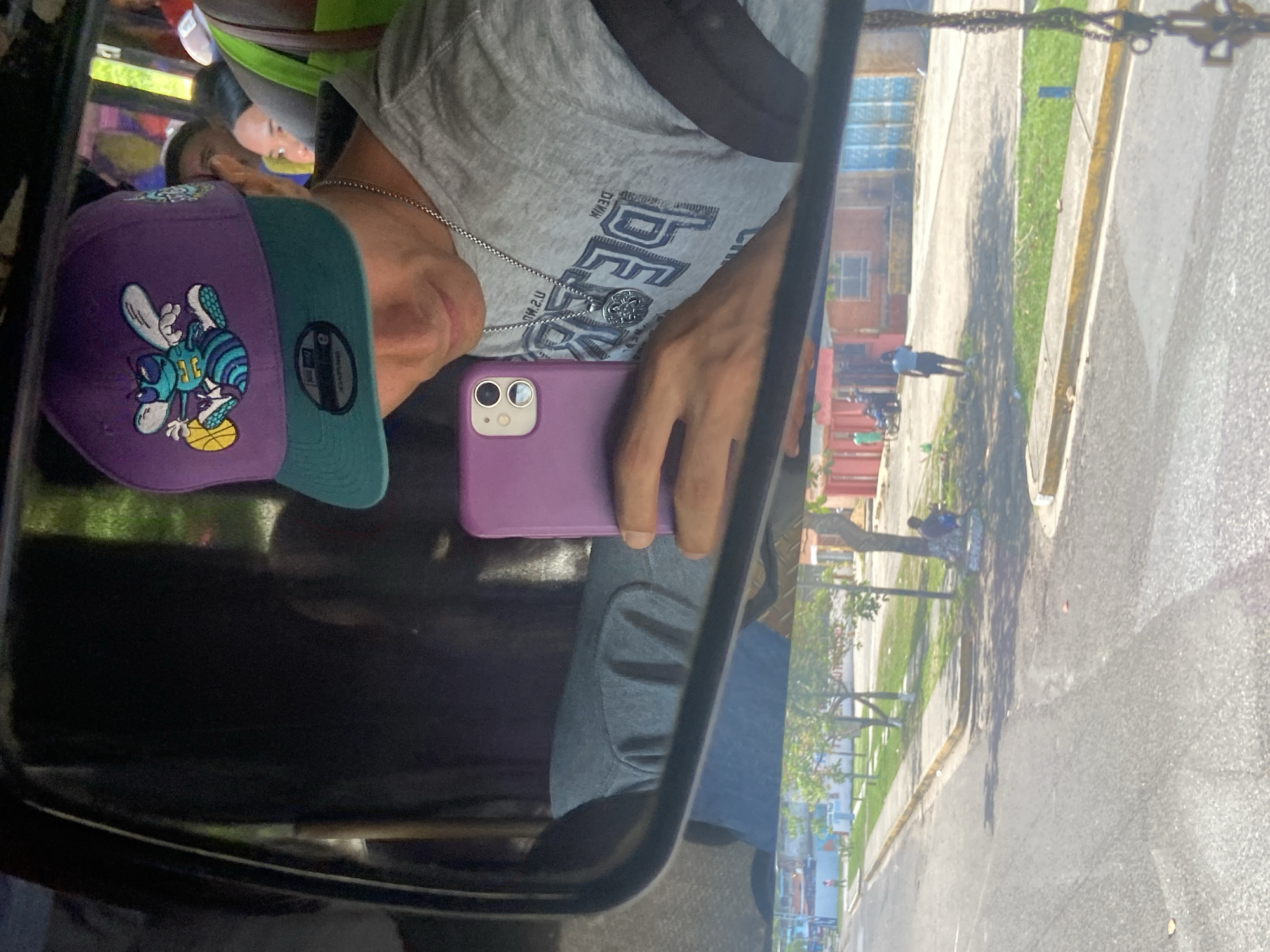 The image captures a selfie in a vehicle mirror, showing a person wearing a grey t-shirt, a purple and green Charlotte Hornets baseball cap, and a silver chain, holding a purple phone. The reflection reveals a street in San José de Barlovento, Venezuela, with buildings, trees, sidewalks, and people walking.