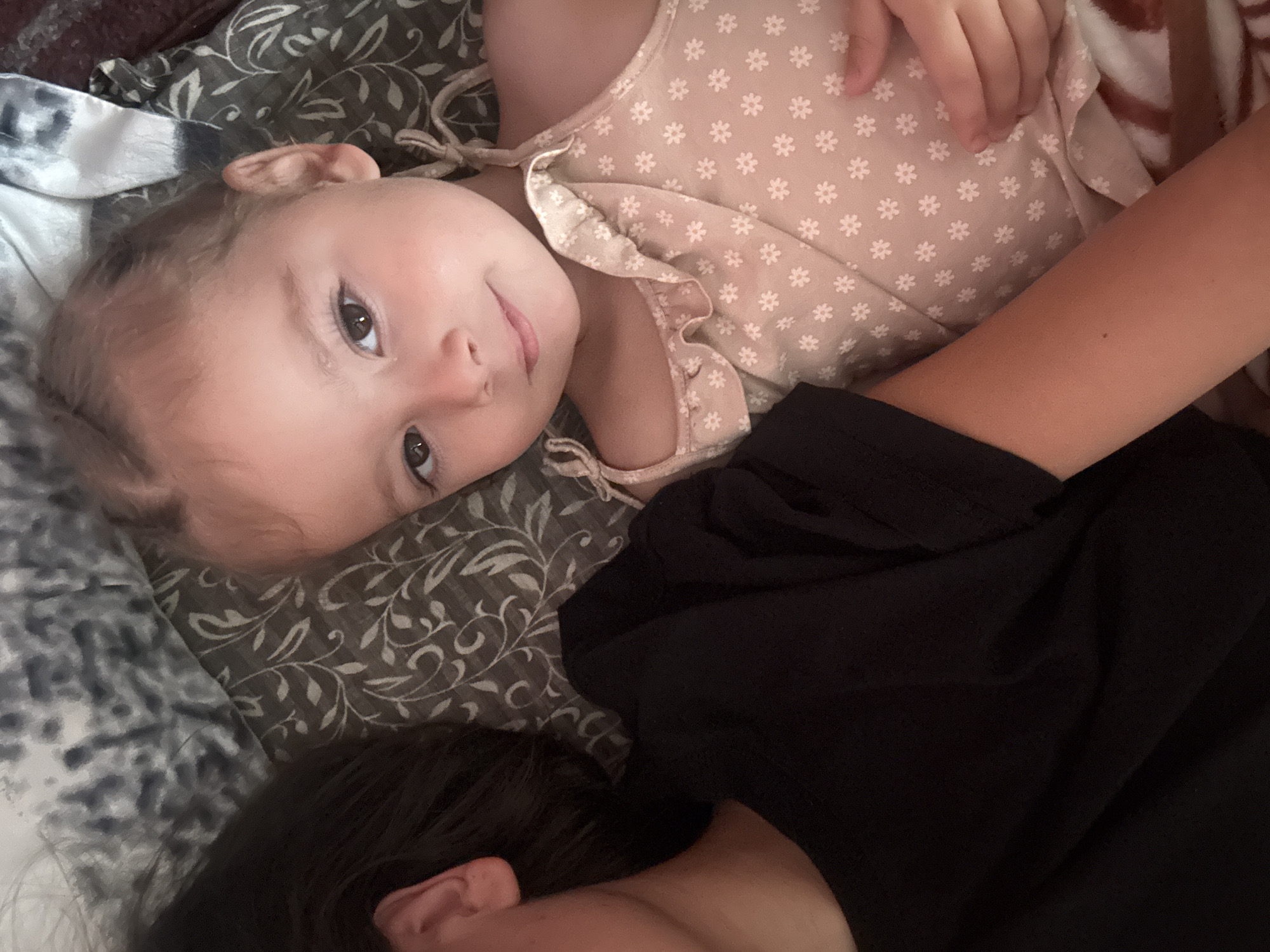 The image shows two individuals lying down, possibly in bed or on a couch. The subject in the foreground is a young child with light brown hair and brown eyes, wearing a light-colored dress with a floral pattern. The child is looking towards the camera with a slight smile. To the right of the child, an arm is visible, and behind them, another person is partially visible, with dark hair and wearing a black shirt. They are lying on a patterned surface, possibly a blanket or duvet, with floral motifs in shades of grey and green. The lighting suggests it might be daytime, but the exact time and weather are not discernible. The overall mood appears relaxed and intimate, possibly indicating a moment of rest or shared quiet time. There is no visible text in the image.