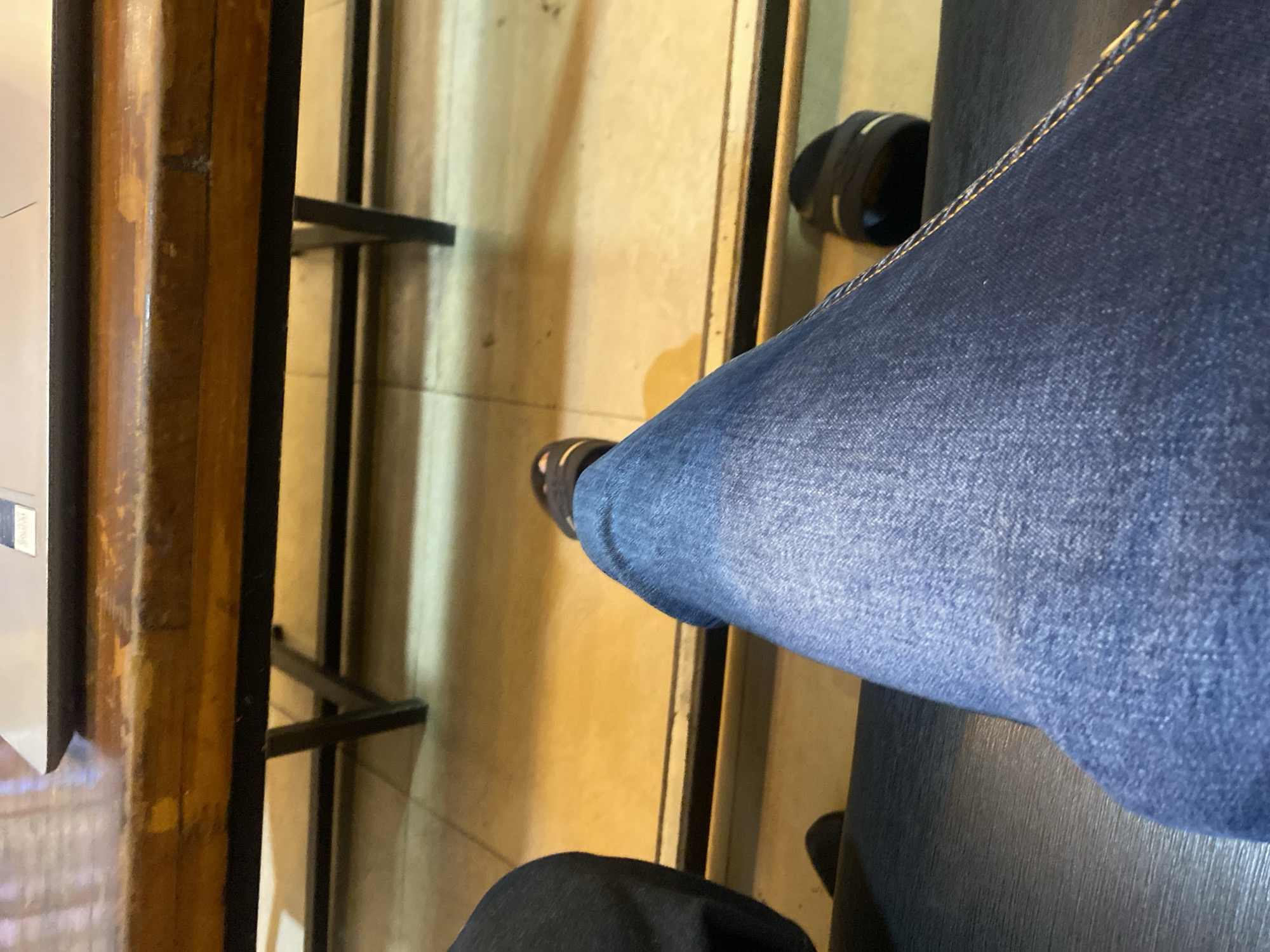 The image captures a low-angle perspective of a person's legs, clad in blue denim jeans, seated at a wooden table. The table has a worn, natural wood finish and is supported by black metal legs, suggesting a rustic or casual setting. The floor beneath the table appears to be tiled in a light, yellowish hue. One foot, wearing a dark sandal, is visible in the foreground. The overall impression is that of a person relaxing or working at a table, possibly in a cafe or outdoor seating area in Lhokseumawe, Indonesia.