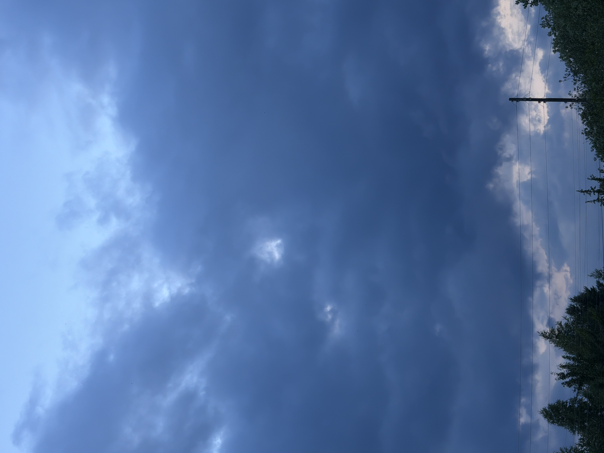 The media file depicts a cloudy sky, viewed from below. The sky is predominantly filled with dark, heavy clouds, suggesting an overcast or impending storm. To the right of the image, a utility pole with power lines stretches vertically, intersecting the cloud cover. Beyond the power lines, a dense canopy of green trees forms a border along the right edge.
The overall impression is one of a somber or dramatic atmosphere, likely due to the dark clouds. The time of day is difficult to ascertain precisely, but the lighting suggests it could be late afternoon or early evening, or a time when the sun is obscured by thick clouds. The presence of trees and utility poles indicates an outdoor, potentially rural or suburban setting, consistent with the provided location context of Petukhi, Russia. No people or specific activities are visible. The image is devoid of any discernible text.