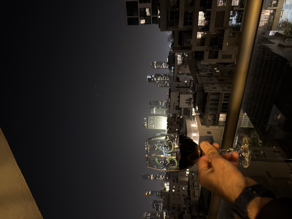 A hand holds a wine glass filled with a dark liquid against a nighttime cityscape. The city features tall buildings, some with illuminated windows, extending into the hazy horizon. A railing is visible in the foreground. The glass has a number "26" written on it in green neon.